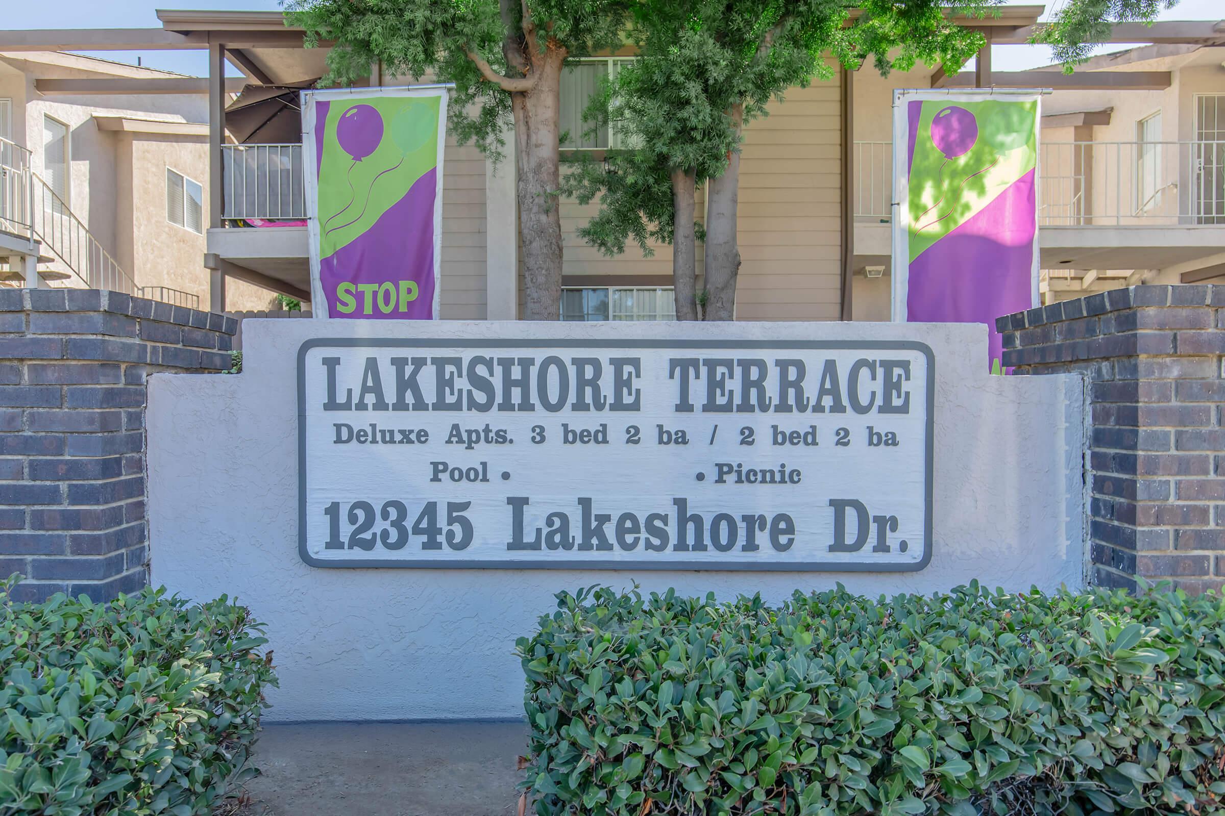 Sign for Lakeshore Terrace apartment community featuring the name "LAKESHORE TERRACE" prominently displayed. Below, it lists available apartments: "Deluxe Apts. 3 bed 2 ba / 2 bed 2 ba." There is a mention of a pool and picnic area. The address "12345 Lakeshore Dr." is also visible, with decorative greenery surrounding the sign.