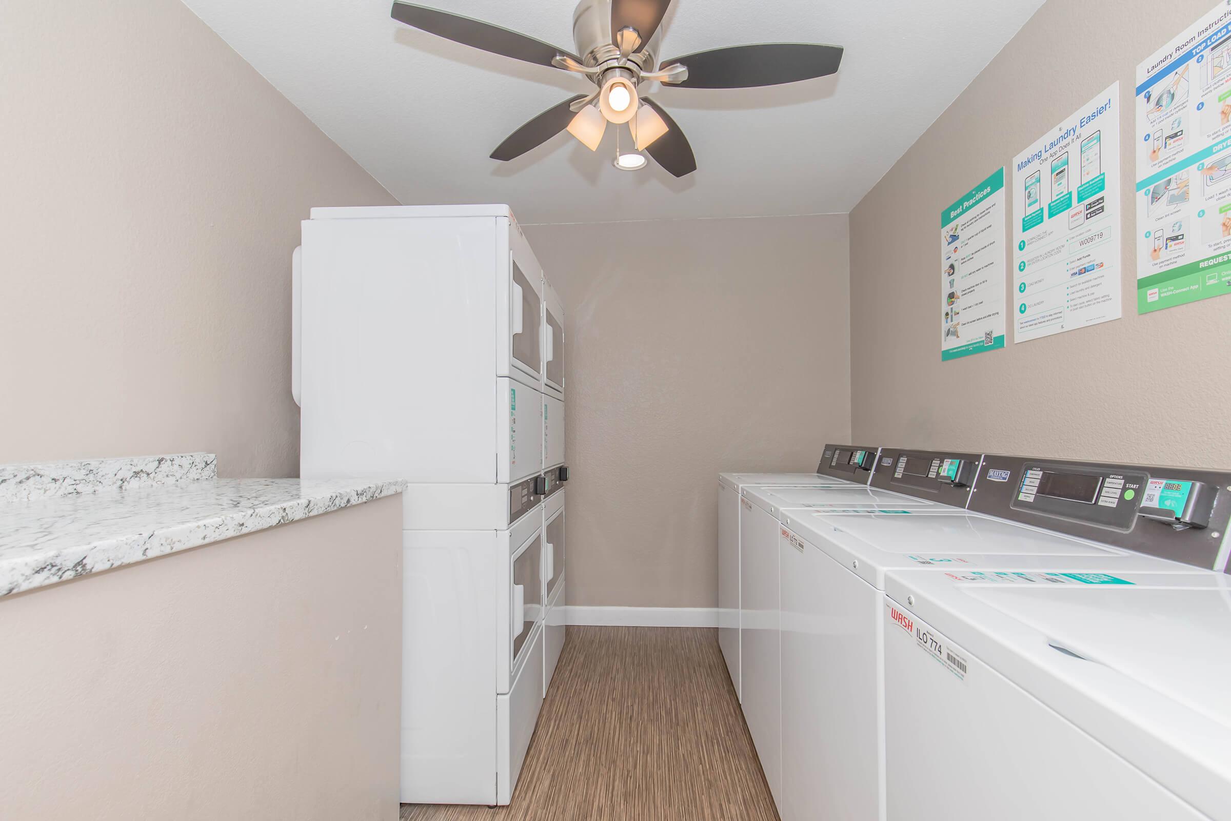 A clean and bright laundry room featuring two sets of stacked washers and dryers alongside a folding counter. A ceiling fan provides ventilation, and informational posters are displayed on the wall. The floor is covered with a wood-like vinyl, creating a modern and inviting atmosphere.