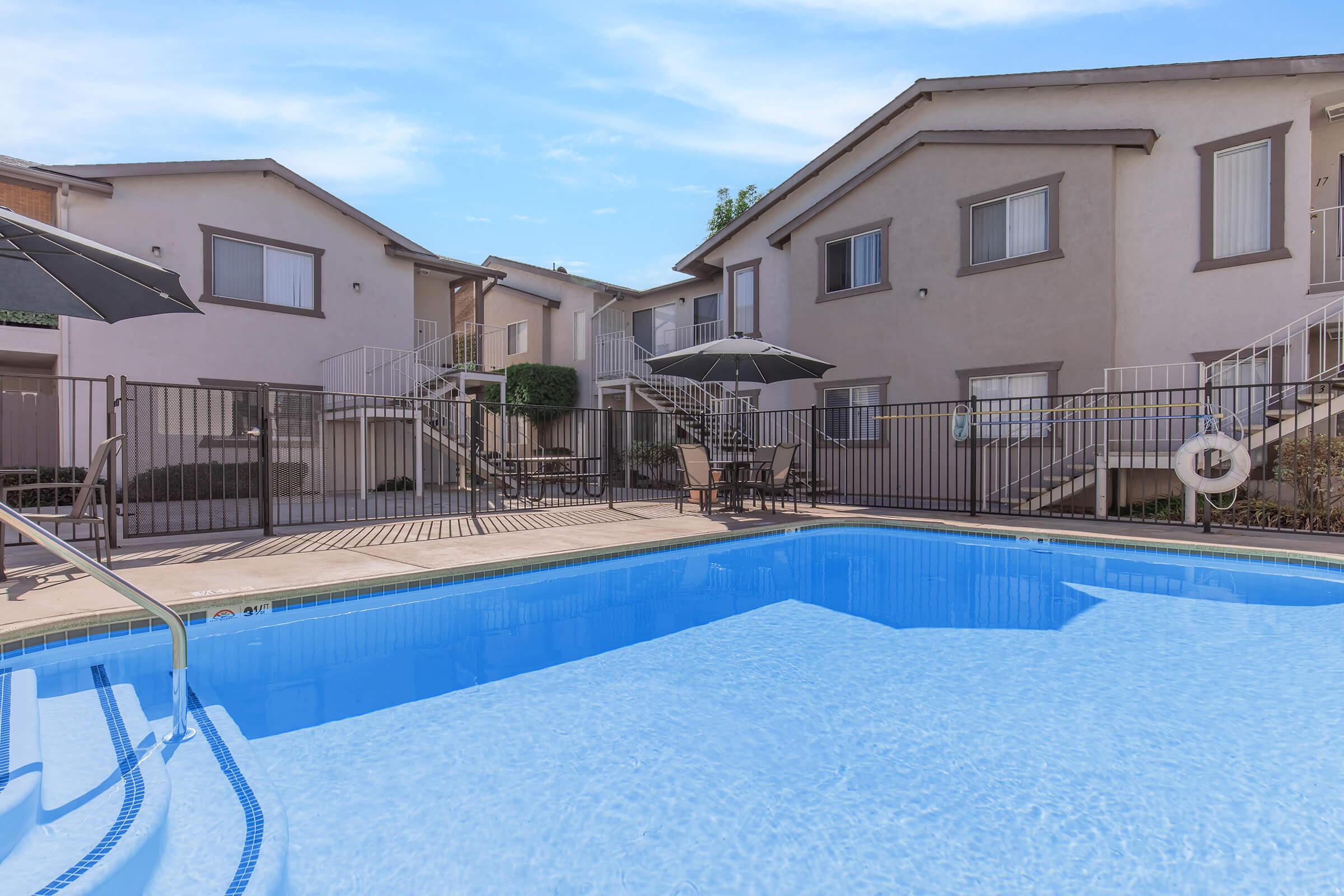 A sparkling blue swimming pool surrounded by a wooden deck and lounge chairs. In the background, two-story apartment buildings are visible, with several umbrellas providing shade. The sky is clear and bright, suggesting a sunny day.