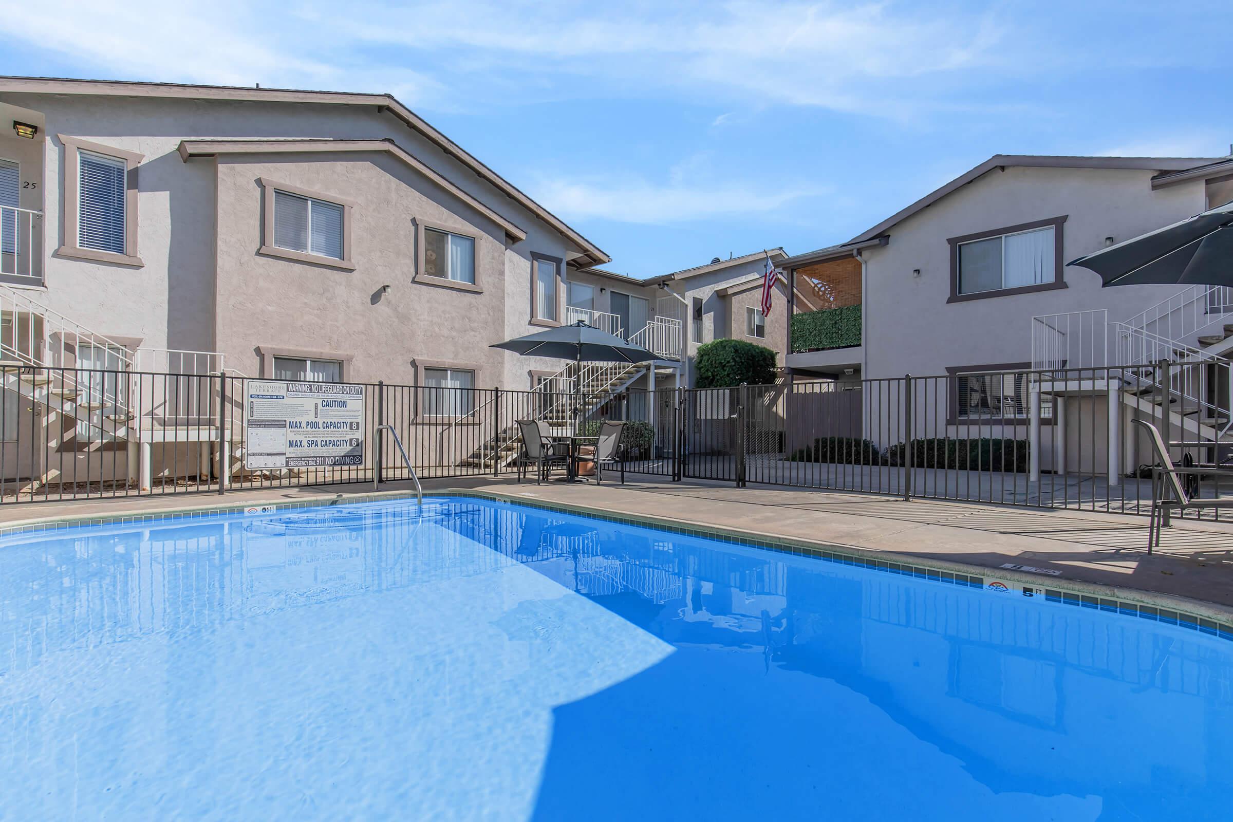 A clear swimming pool in the foreground with apartment buildings surrounding it. The buildings feature balconies and outdoor seating areas, with umbrellas for shade. The scene is bright and sunny, showcasing a well-maintained outdoor space ideal for relaxation and leisure.