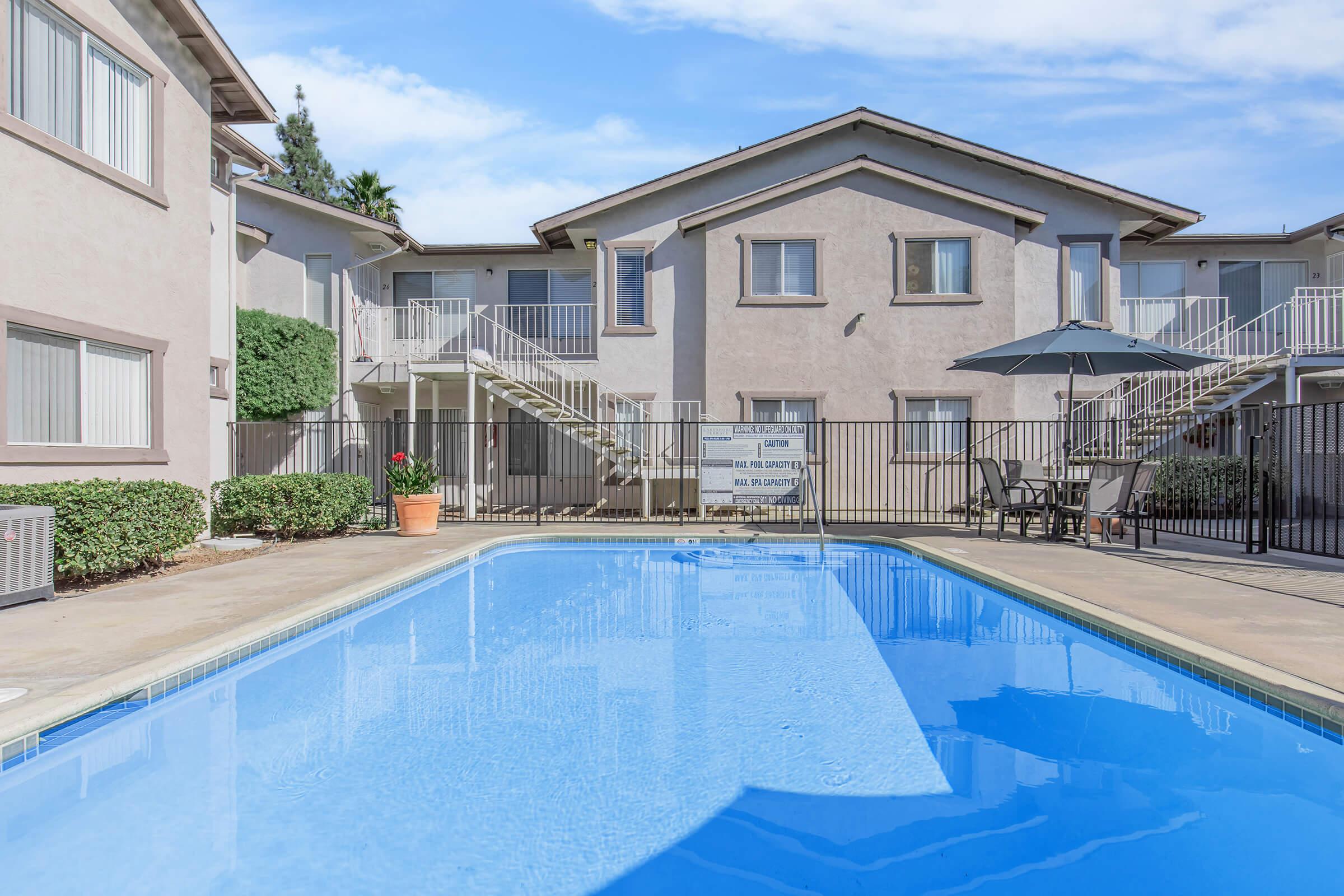 A clear blue swimming pool surrounded by a well-maintained outdoor area, including two buildings with balconies. There are tables and chairs under an umbrella near the pool, and lush greenery on the sides. The sky is bright with a few clouds, creating a inviting atmosphere.