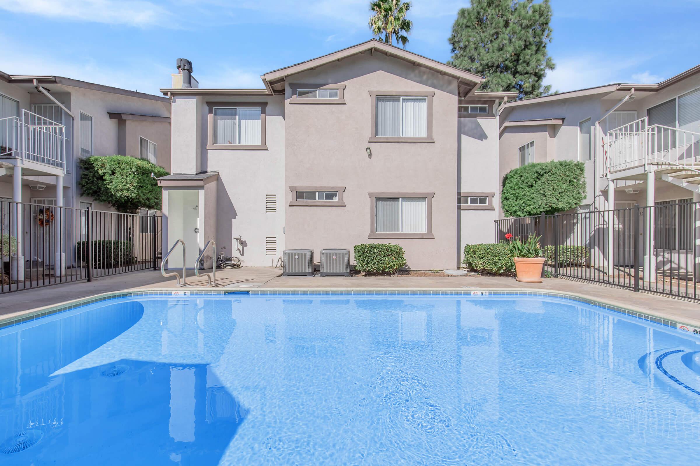 A well-maintained swimming pool in front of a multi-story apartment building. The pool reflects the blue sky and is surrounded by a clean patio, with well-trimmed shrubs and a potted plant nearby. The building features several windows and a balcony, adding to the inviting atmosphere of the area.