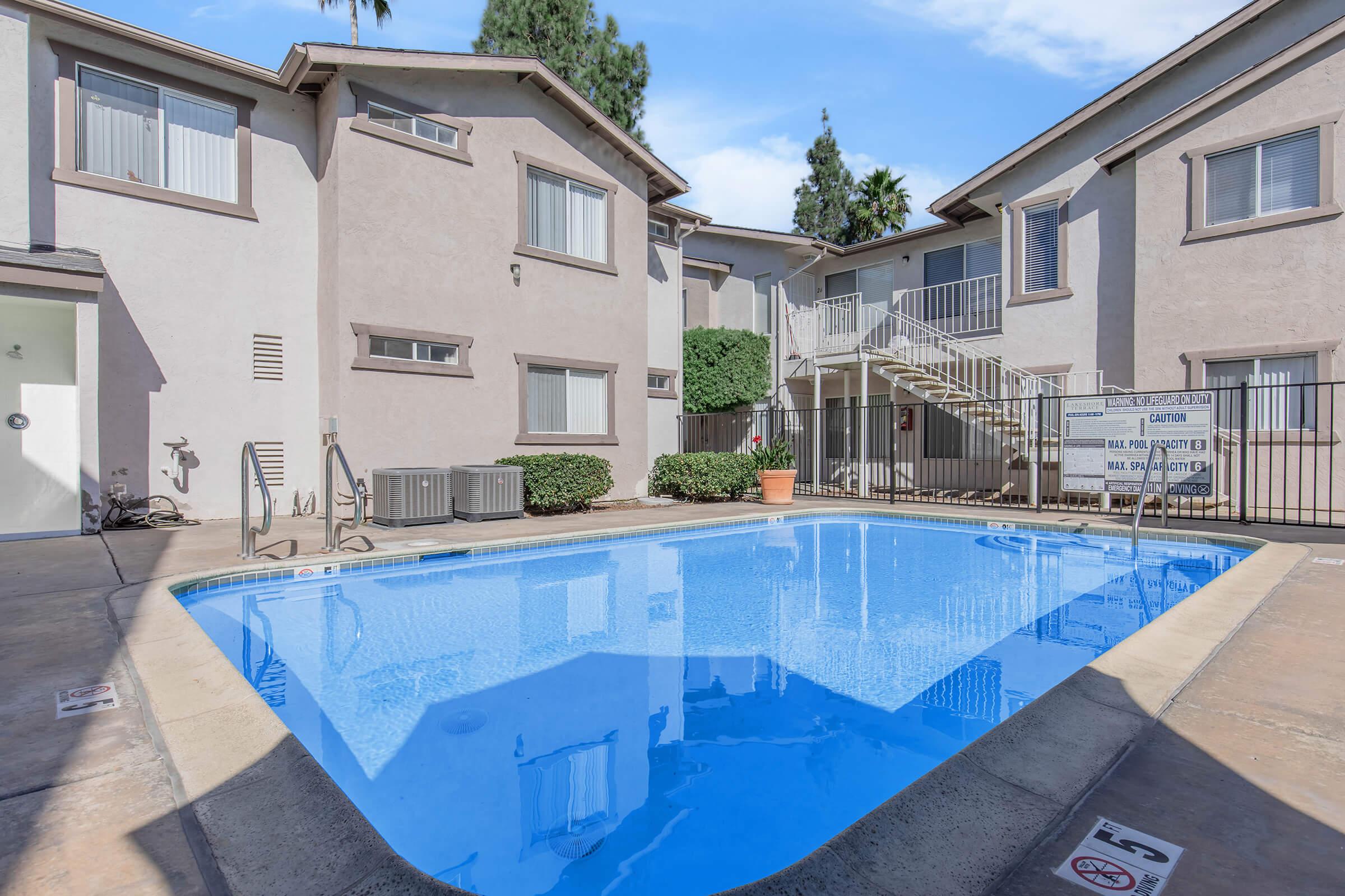 A clean, blue swimming pool surrounded by beige apartment buildings. The pool area features a concrete deck, potted plants, and a staircase leading to the upper level of the apartments. Windows of the apartments overlook the pool, creating a cozy outdoor space for residents.