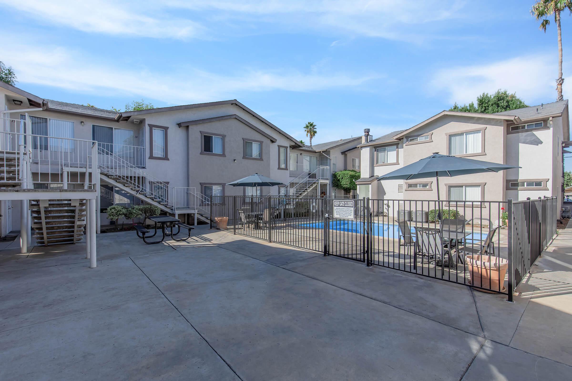 A sunny courtyard of an apartment community featuring multiple two-story buildings. A swimming pool is enclosed by a fence, surrounded by lounge chairs and umbrellas. There are picnic tables and stairs leading to upper-level units, with clear blue skies and palm trees in the background.