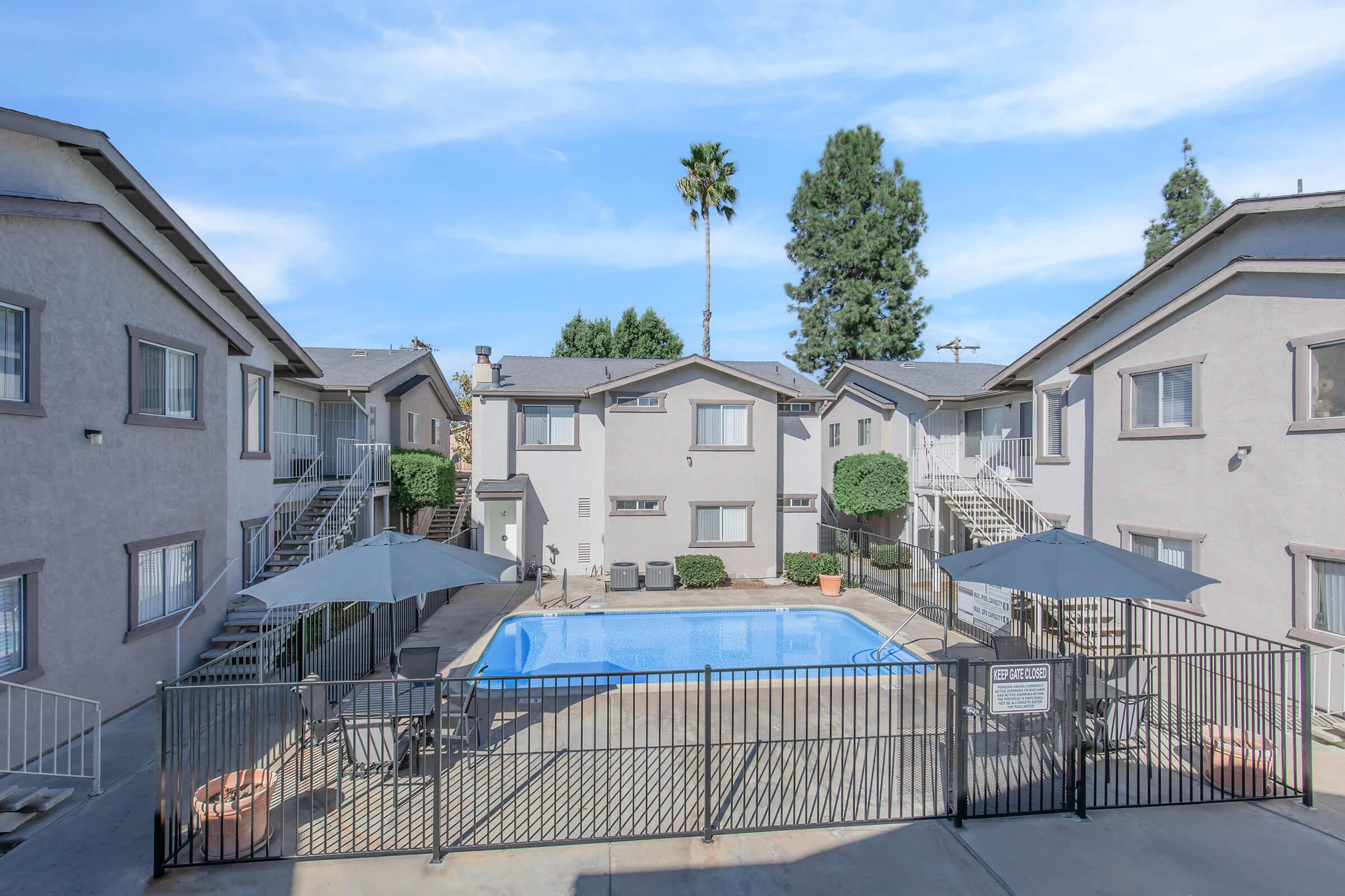 A view of an apartment community featuring a swimming pool surrounded by a gated area. The buildings are two stories high, with staircases leading up to the apartments. Shade umbrellas and lounge chairs are placed around the pool area, with trees and greenery in the background under a clear blue sky.
