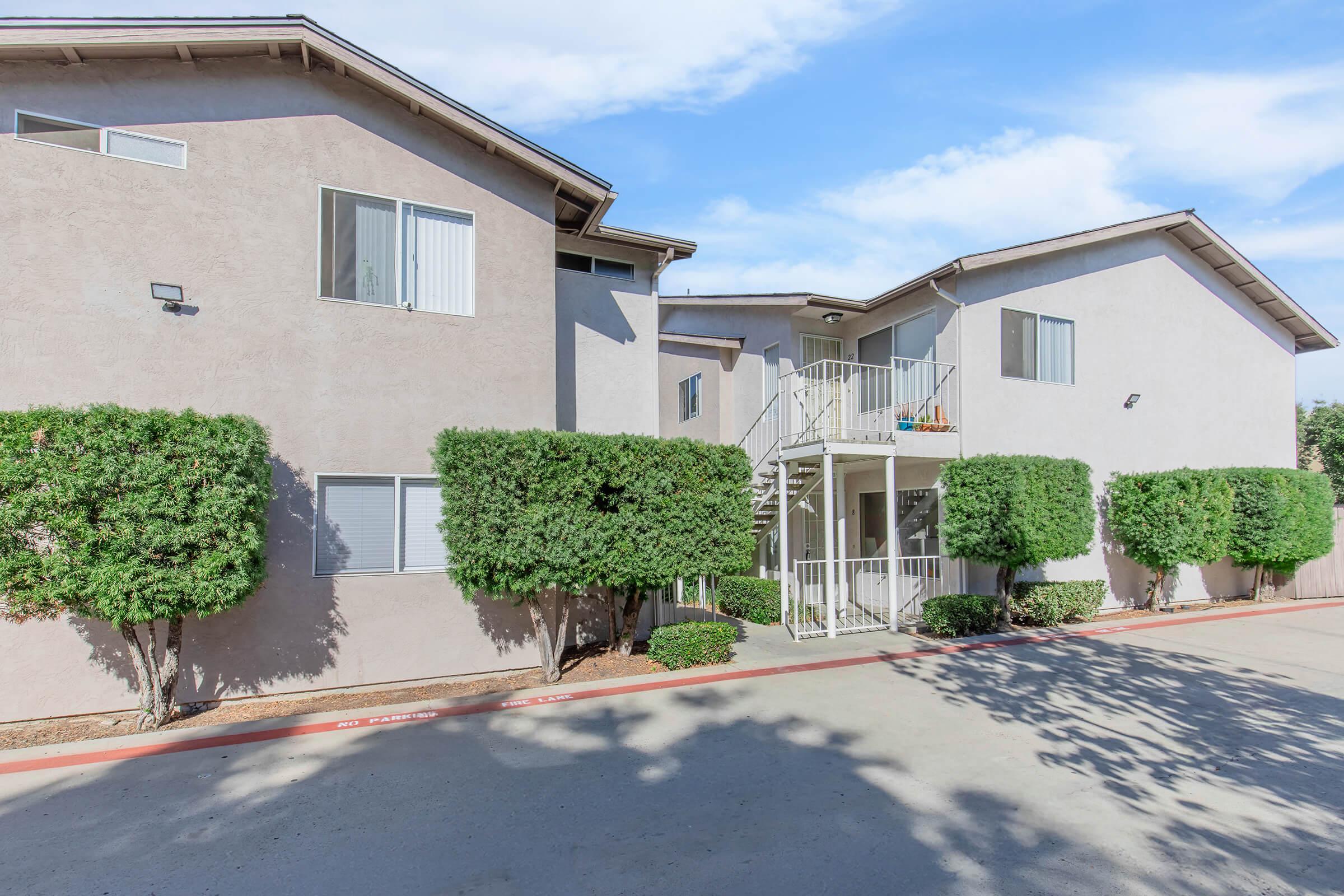 A two-story residential building with a light gray exterior. The building features several windows, a staircase leading to a second-floor entrance, and neatly trimmed green bushes in front. The surrounding area has a paved driveway, with blue skies and clouds in the background.