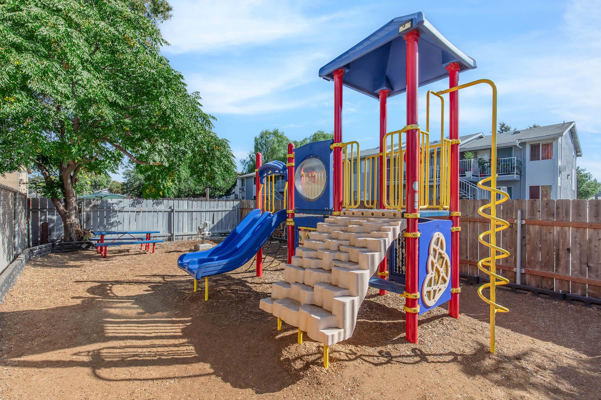 A colorful playground featuring a climbing structure with slides, stairs, and safety rails, surrounded by soft earth. The area is enclosed by a wooden fence, with benches and picnic tables in the background under a blue sky and large trees.