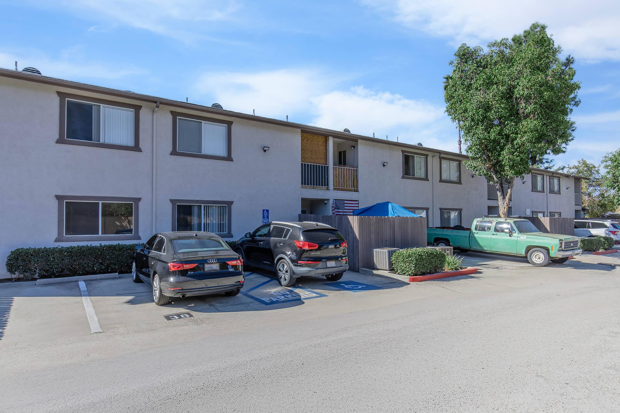 A parking area in front of a two-story apartment building. The building features several windows and a balcony. Two modern black cars are parked in designated spots, while a vintage green pickup truck is parked alongside. A shaded area with a blue tarp is visible, and there is a well-maintained hedge surrounding the premises.