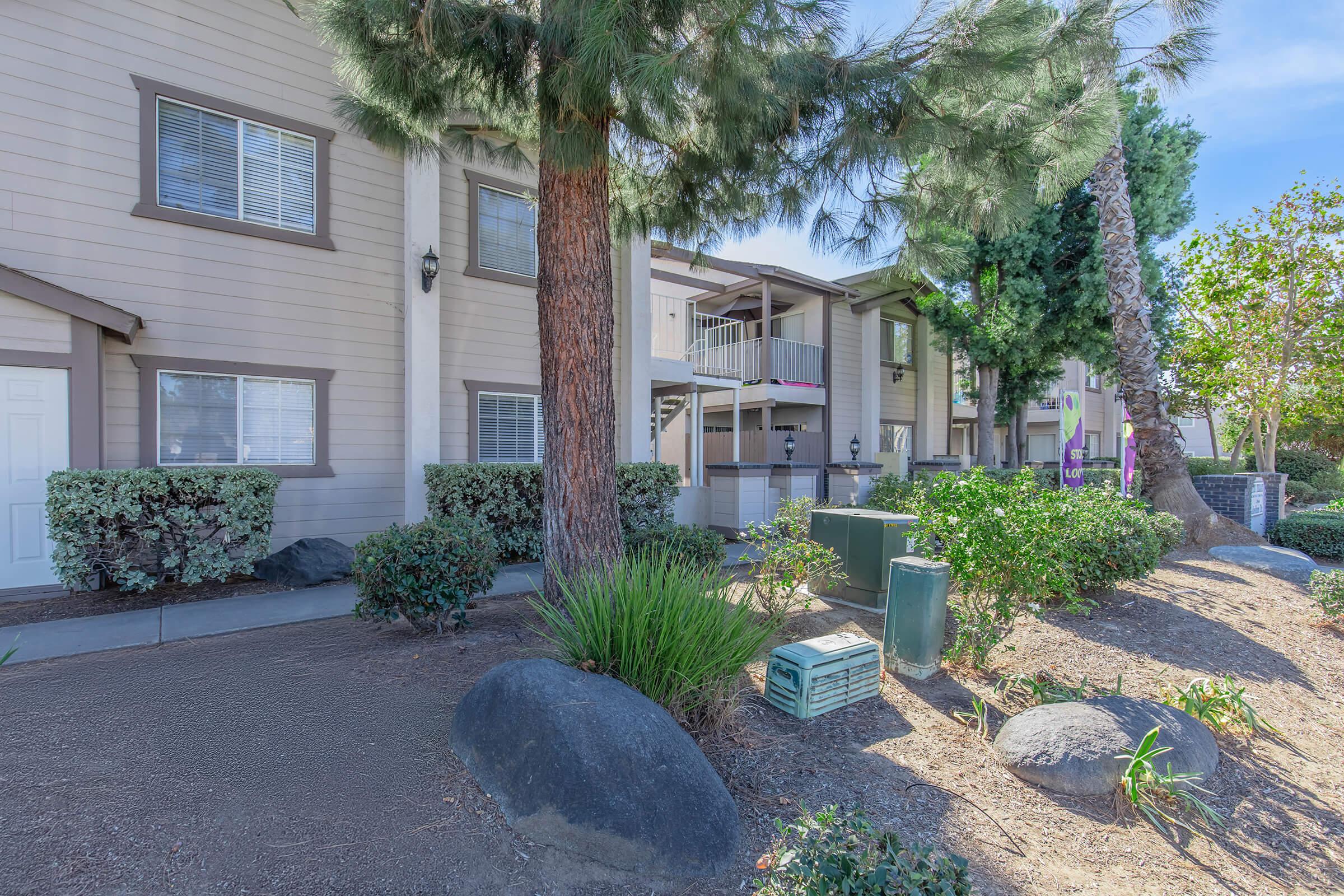 Residential apartment community exterior showing multiple stories, surrounded by greenery and trees. There are shrubs and a lawn area, along with utility boxes visible on the ground. The building features windows and balconies, with a clear blue sky in the background.