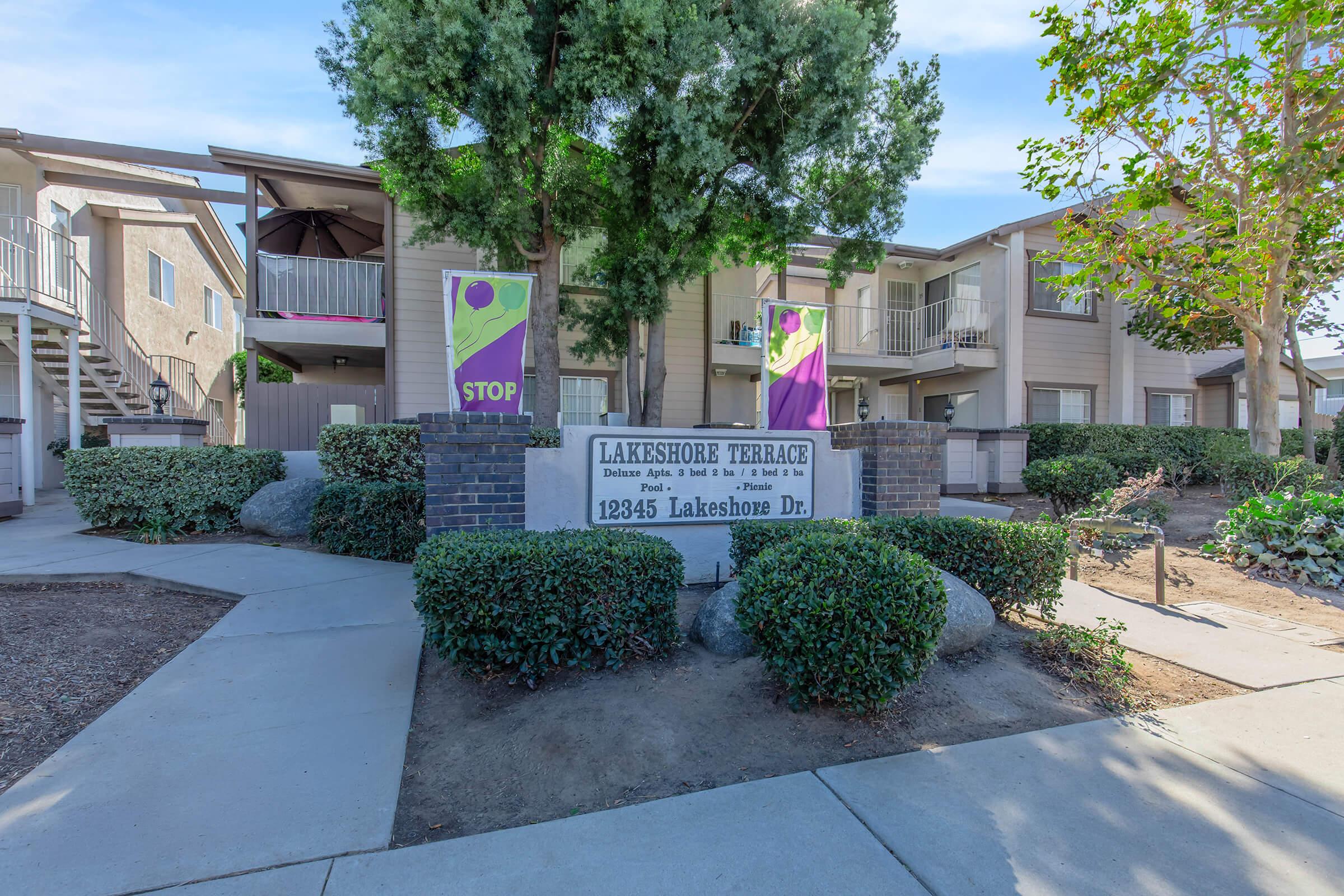 Lakeshore Terrace apartment community entrance featuring a sign with the address "12345 Lakeshore Dr." Surrounded by landscaped bushes and trees, two banners are visible, one displaying a stop sign. The building has a two-story design with staircases leading to upper units.