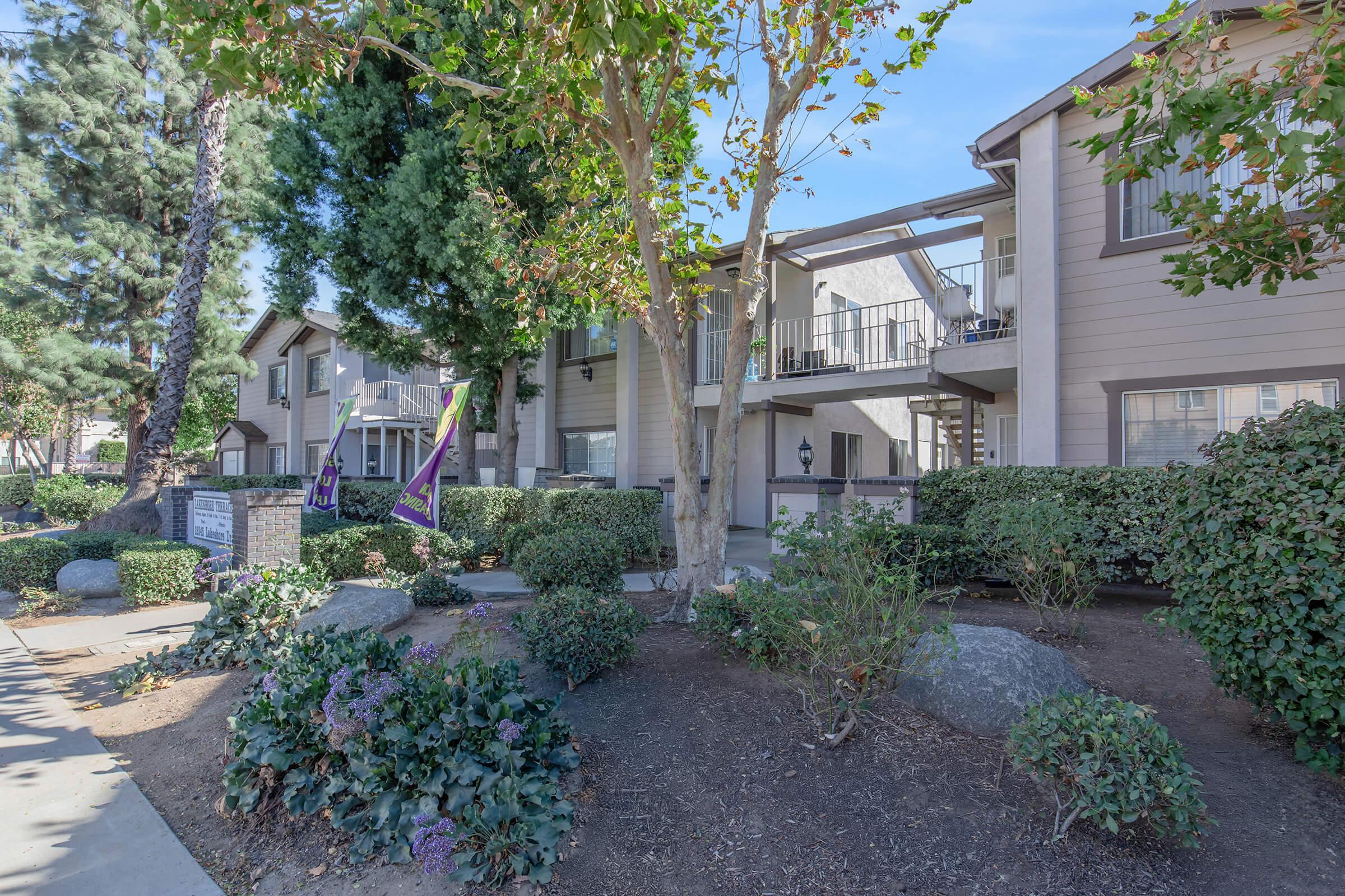 A landscaped apartment community with two-story buildings surrounded by shrubs and trees. There are stone pathways, greenery, and decorative rocks in the foreground. The sky is clear and blue, adding to the inviting atmosphere.
