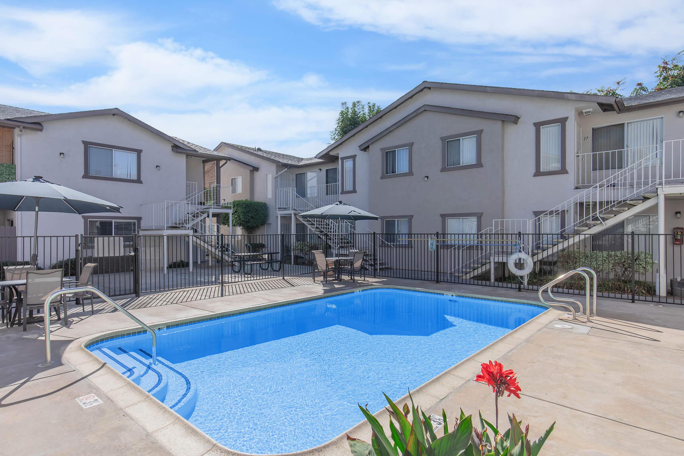 A tranquil courtyard featuring a sparkling blue swimming pool surrounded by lounge chairs and small tables. In the background, there are two stories of apartment buildings with staircases leading to the upper level. Decorative plants and umbrellas create a relaxing outdoor atmosphere under a bright blue sky.