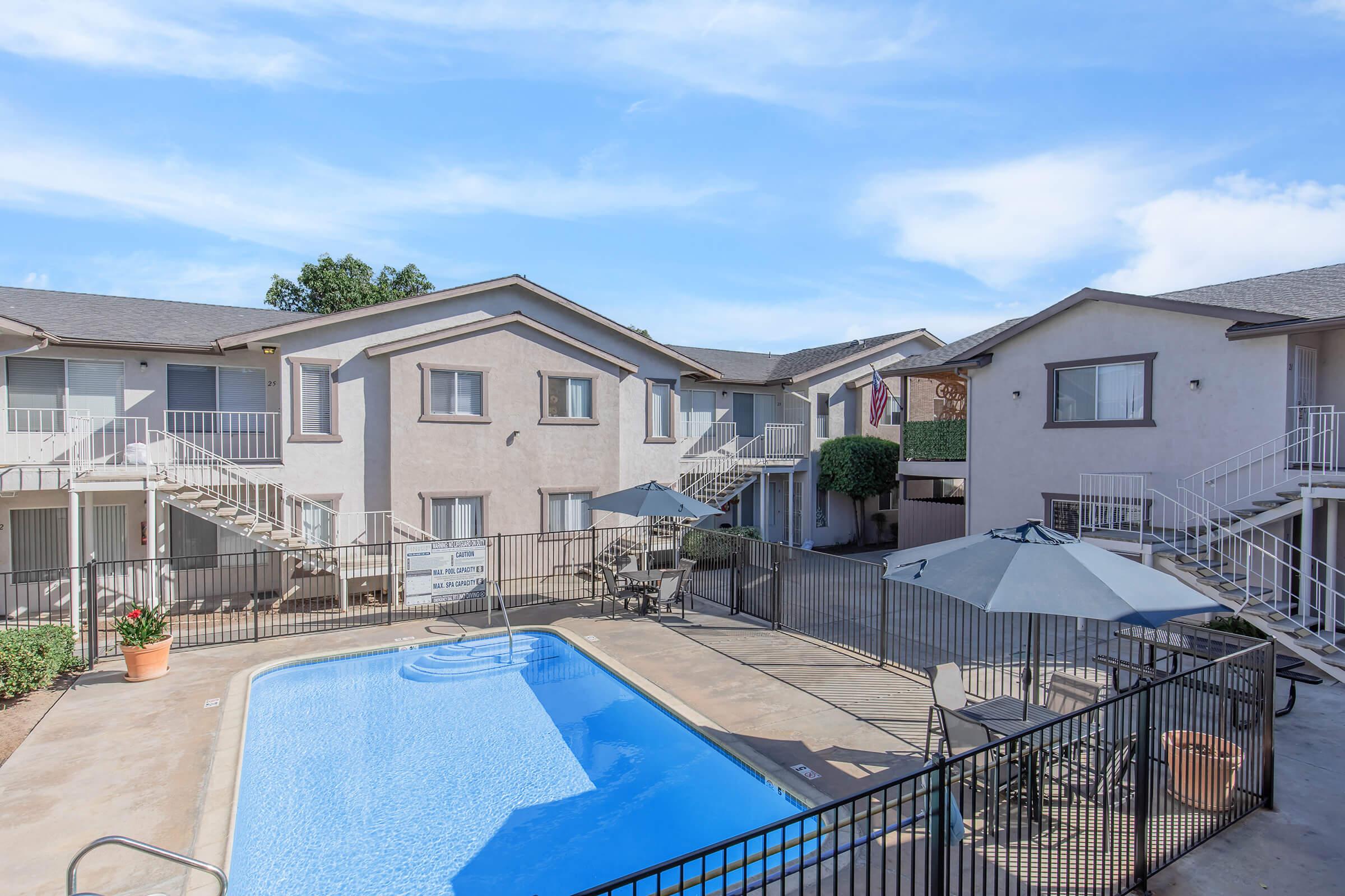 A view of an apartment community featuring a swimming pool at the center. Surrounding the pool are lounge chairs and tables with umbrellas, along with well-maintained landscaping. The buildings have balconies and stairs, with a clear blue sky in the background.