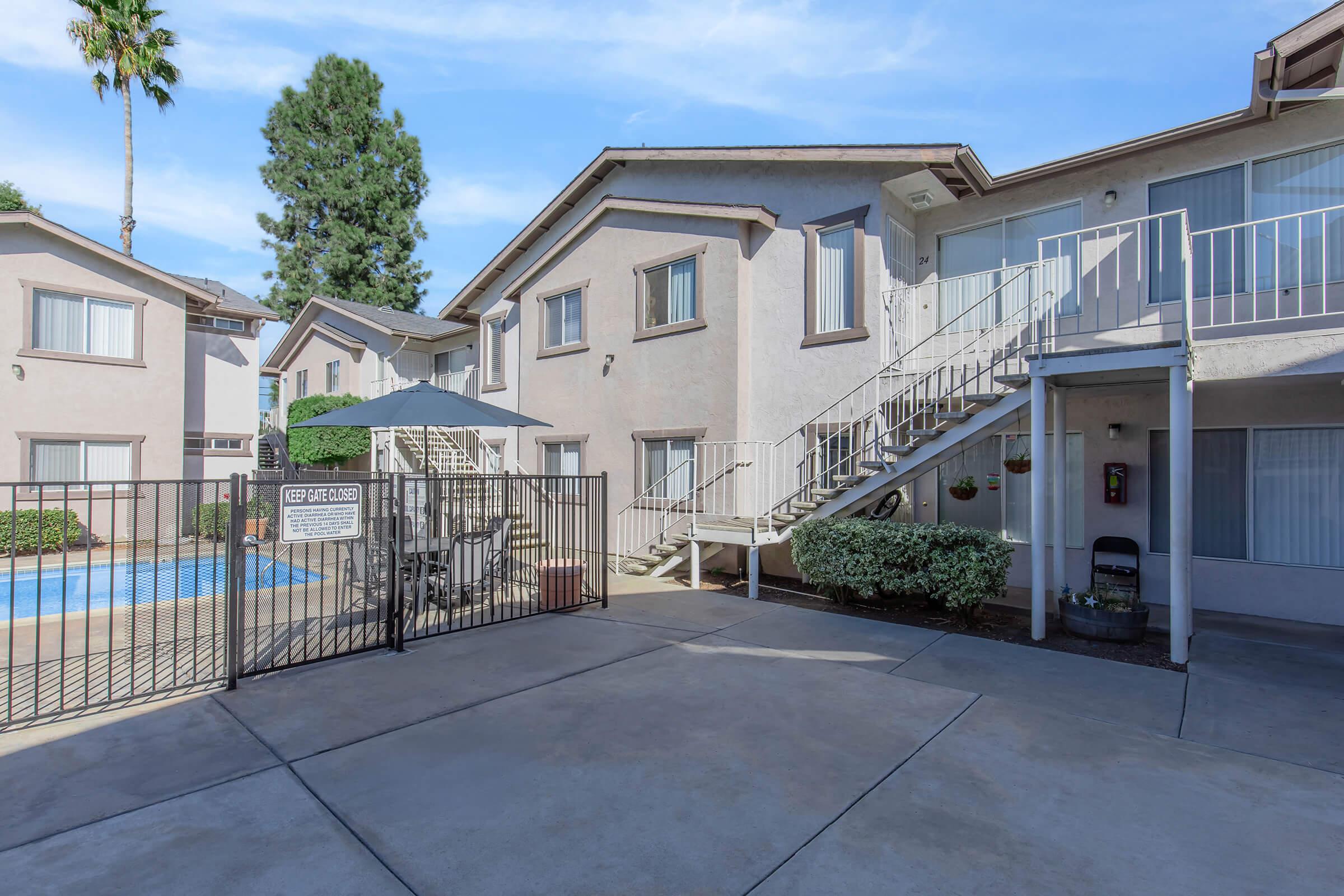 A view of an apartment community featuring a swimming pool area surrounded by a gated fence. The buildings are two stories high, with a staircase leading to the second floor. There are palm trees in the background and a shaded seating area nearby, creating a welcoming outdoor space.