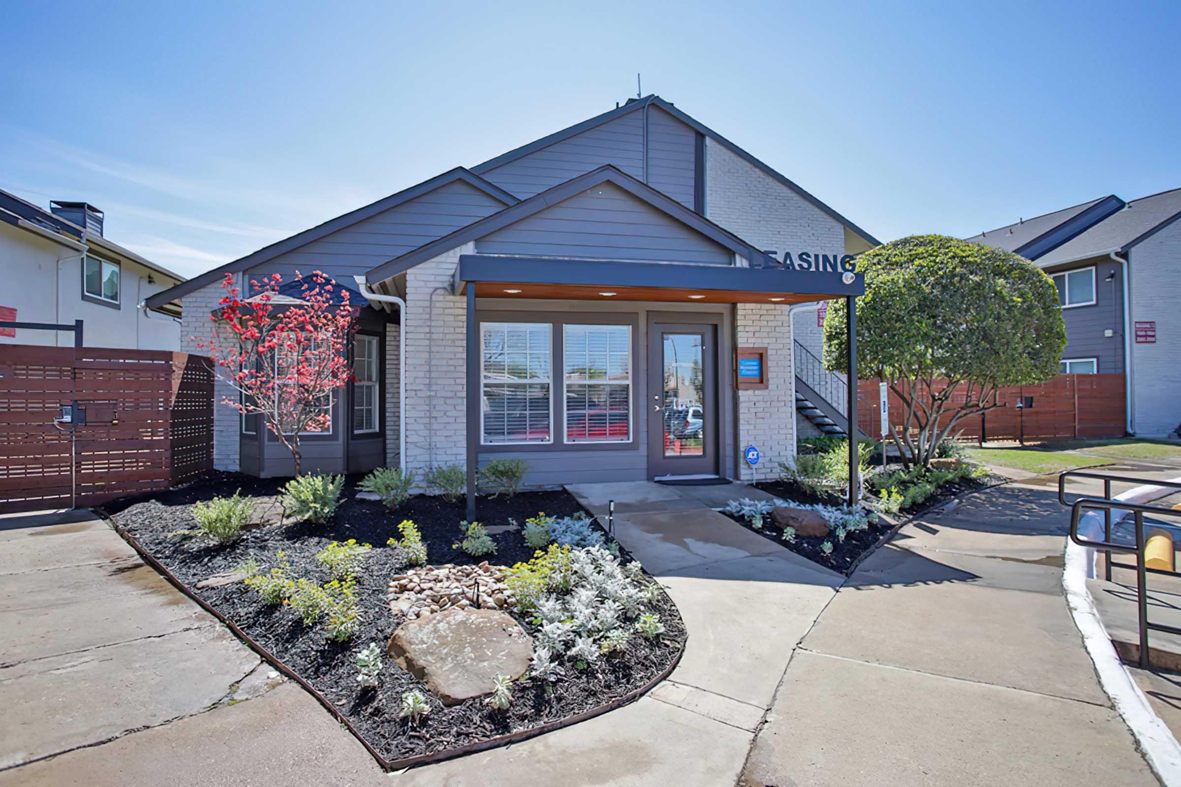 A modern leasing office with a welcoming entrance. The building features gray siding and a peaked roof, surrounded by landscaped gardens with colorful flowers and shrubs. A pathway leads to the front door, and a sign indicates the leasing office. Clear blue skies enhance the bright and inviting atmosphere.