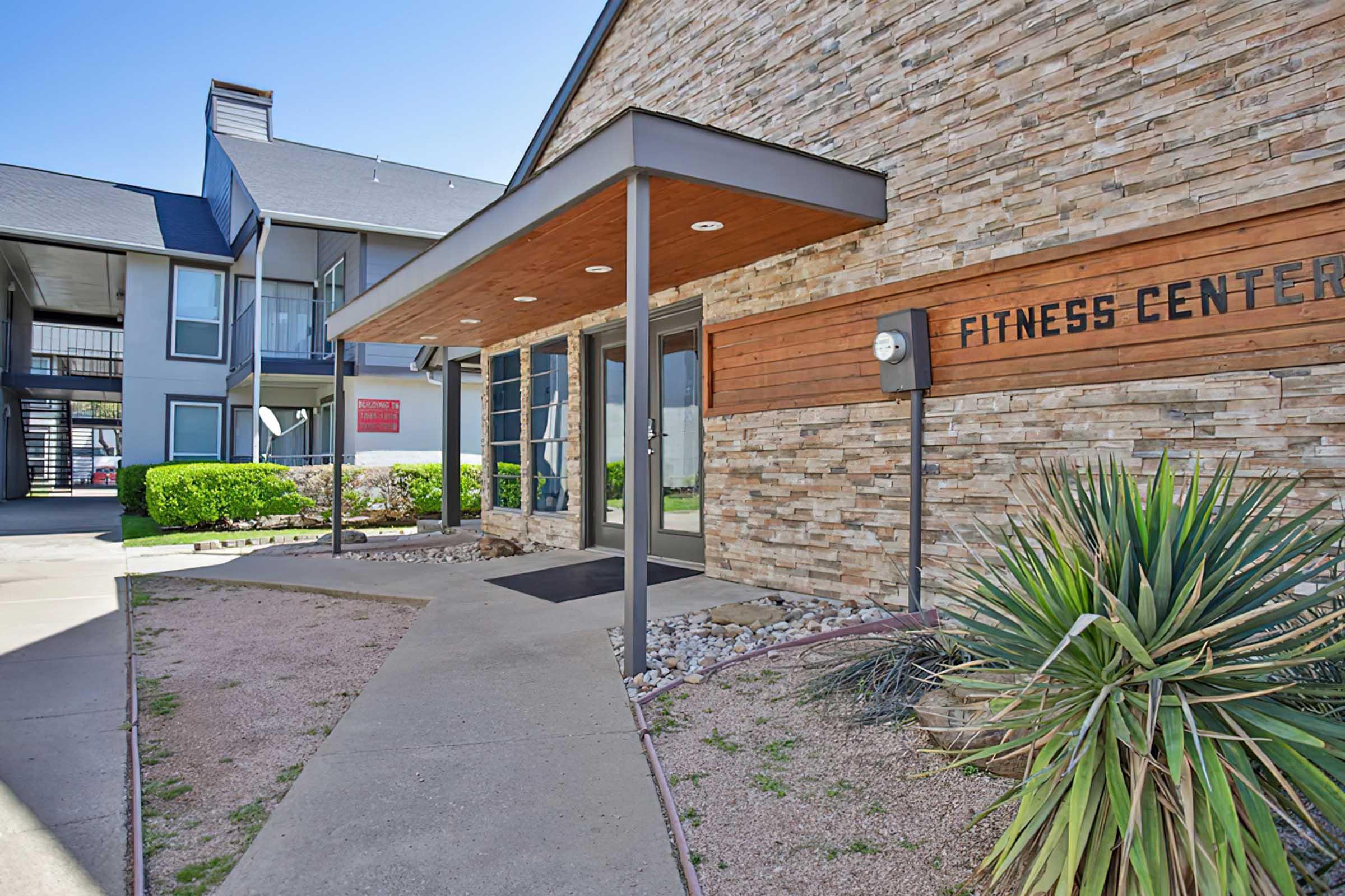 Entrance to a fitness center with a modern design, featuring a stone facade and wooden accents. The area is landscaped with shrubs and rocks, and there are nearby buildings visible in the background. The fitness center sign is prominently displayed on the wall.