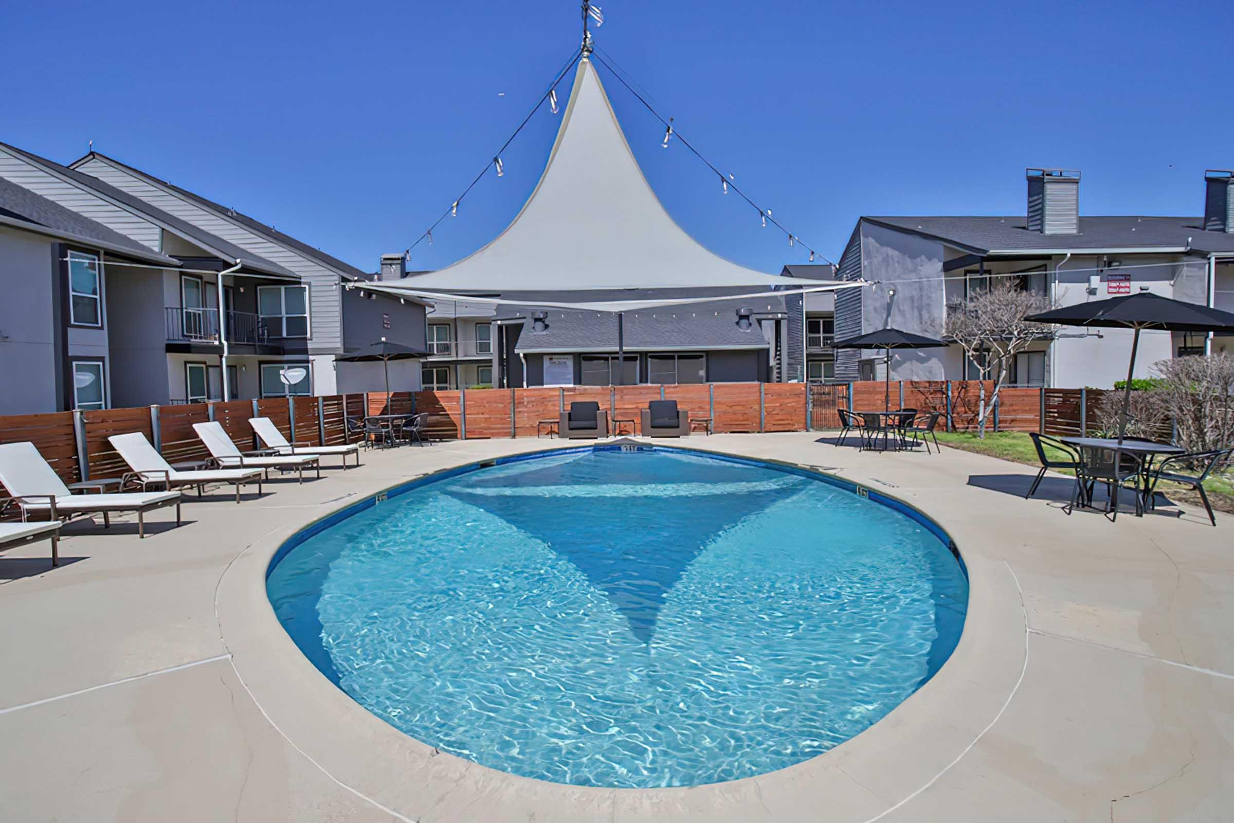 A clear blue swimming pool surrounded by lounge chairs and patio tables. The pool area features a large white canopy overhead and is set within an apartment complex, with buildings visible in the background. Bright sunlight creates a cheerful atmosphere.