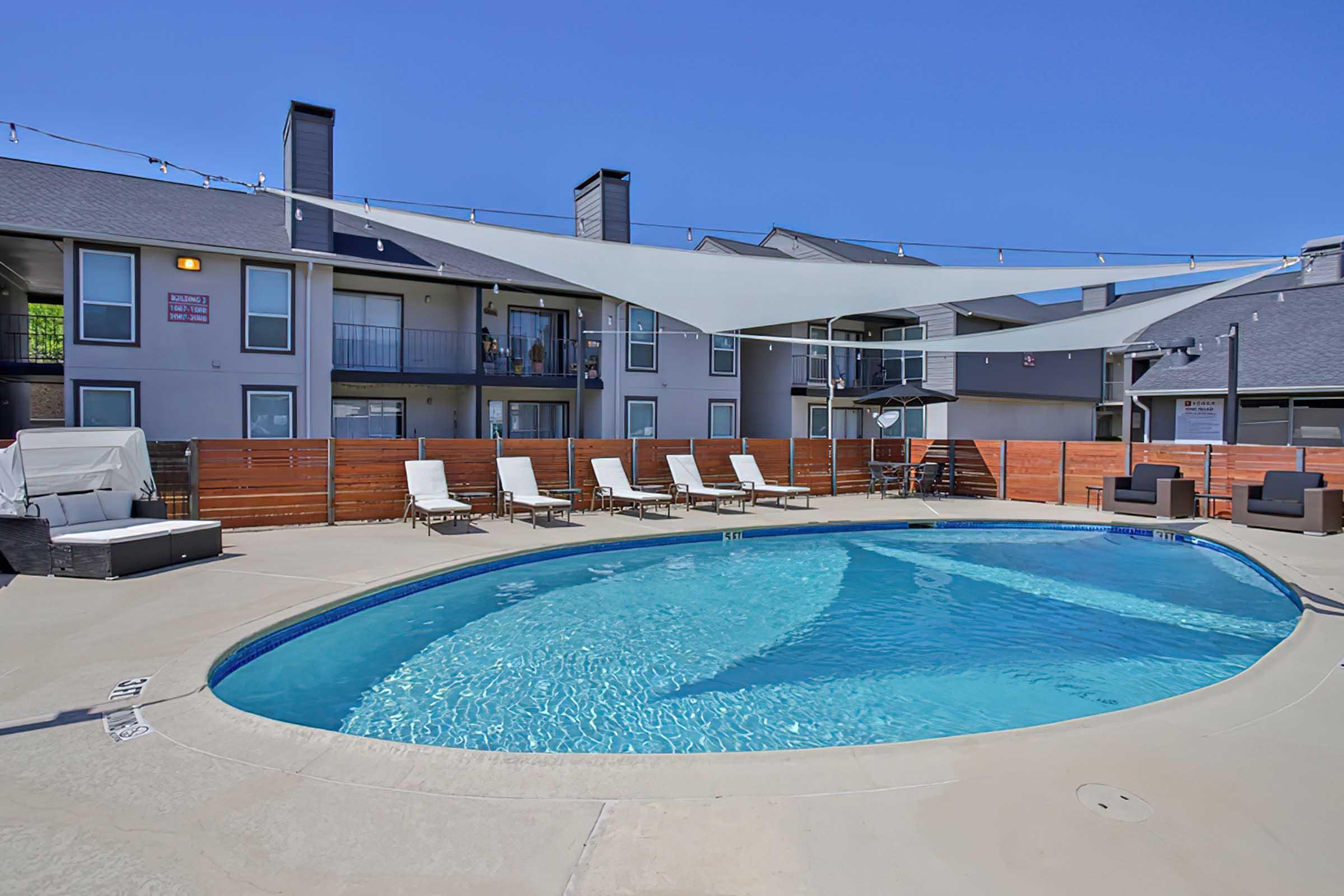 A swimming pool surrounded by lounge chairs and shaded areas. In the background, there are multiple apartment units with balconies. The sky is clear and blue, creating a bright, inviting atmosphere.
