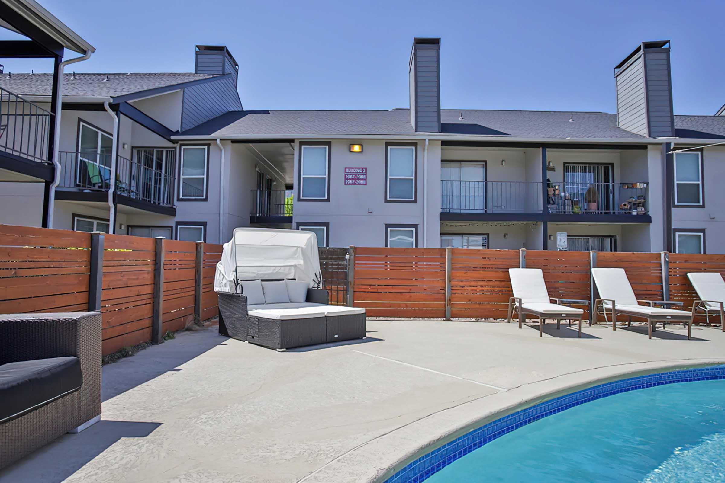 Poolside area featuring a comfortable lounge chair and a shaded seating area. In the background, there are balconies of an apartment building, and the pool is visible with a curved edge. The surrounding fence is made of wooden panels, creating a cozy outdoor atmosphere. Clear blue skies above.