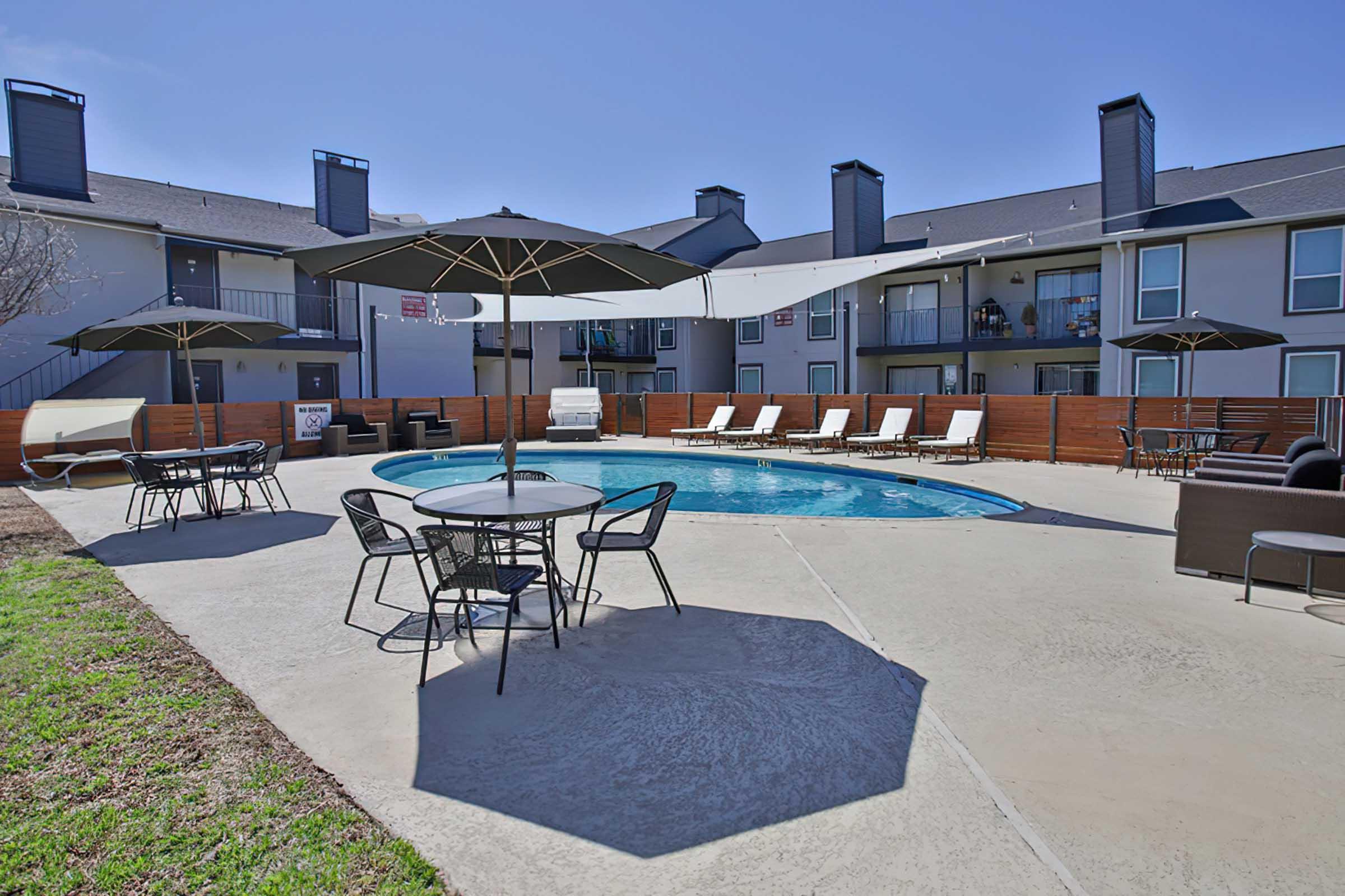 A sunlit outdoor pool area featuring a rectangular swimming pool surrounded by lounge chairs and tables. Large umbrellas provide shade, with a grassy area nearby. Apartment buildings are visible in the background, creating a relaxed residential atmosphere.
