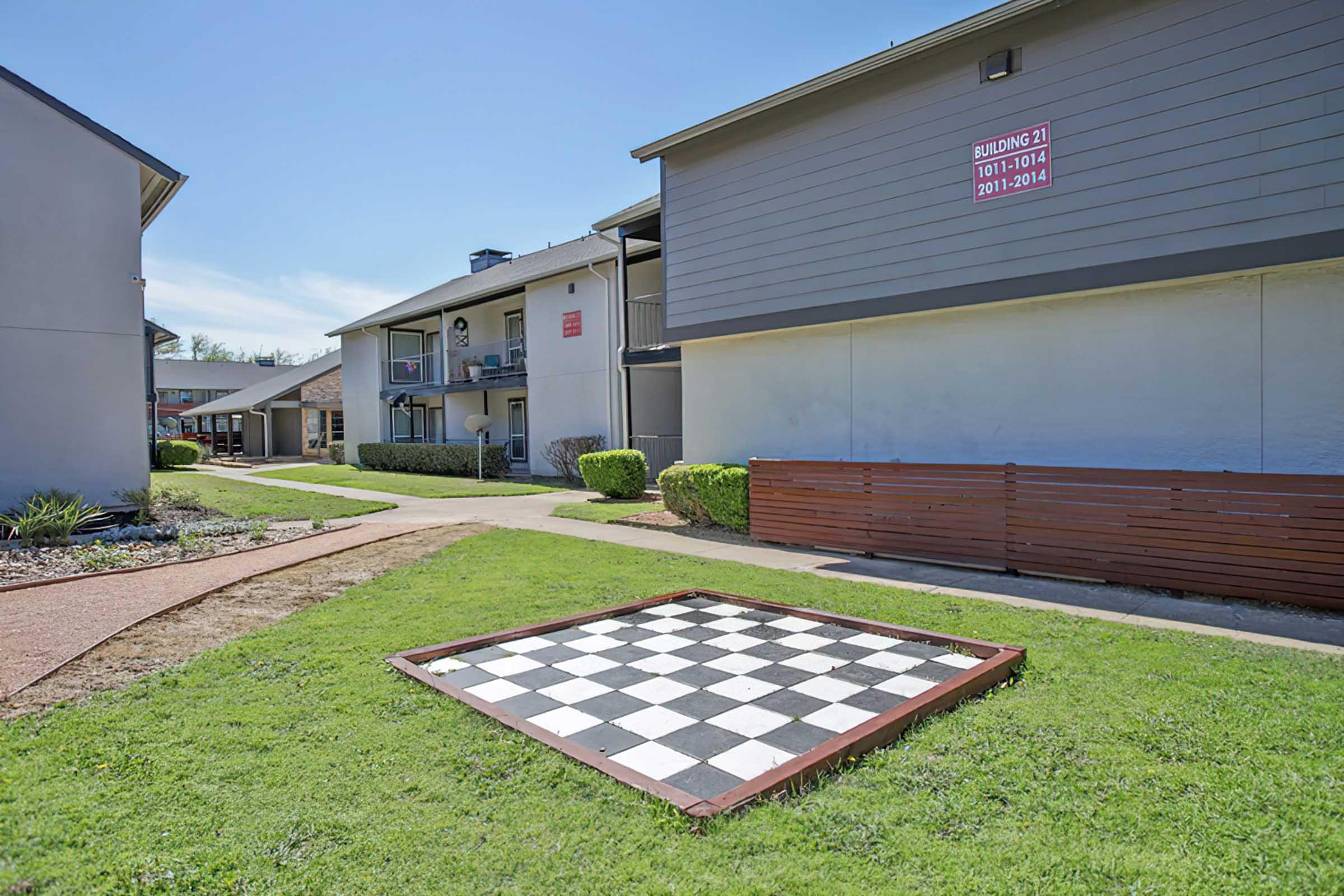 A large outdoor chessboard made of black and white squares on a green lawn, bordered by small shrubs. In the background, there are apartment buildings with balconies, and a clear blue sky overhead. The scene is well-maintained and inviting, perfect for outdoor activities.