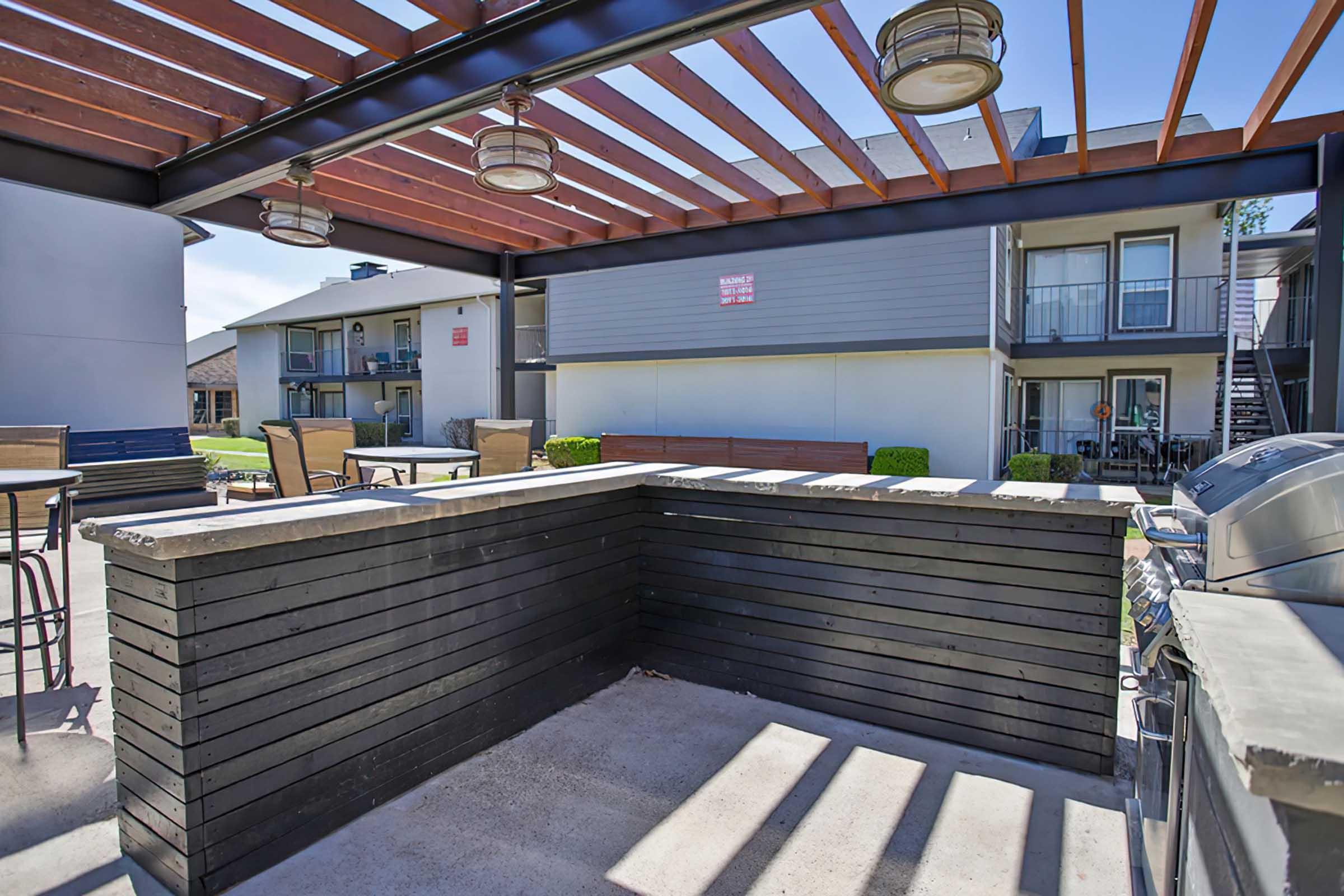 An outdoor patio area featuring a wooden bar with seating, shaded by a slatted roof. In the background, there are residential buildings with balconies. The setting is bright and sunny, perfect for social gatherings or relaxation. A grill is visible near the bar area.