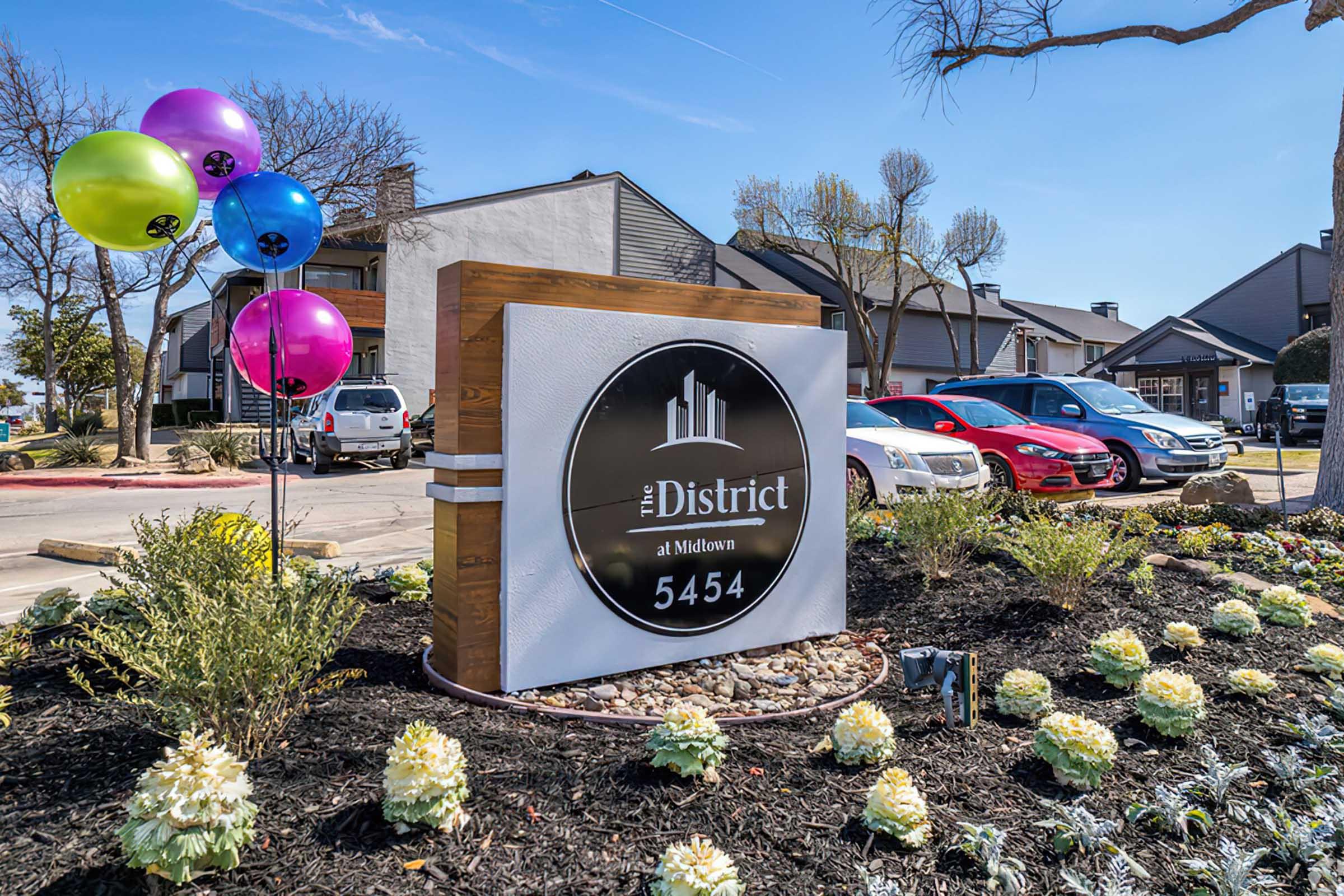 Sign for "The District at Midtown" featuring the address 5454, surrounded by colorful balloons and landscaped plants. In the background, several cars are parked, and there are residential buildings visible.