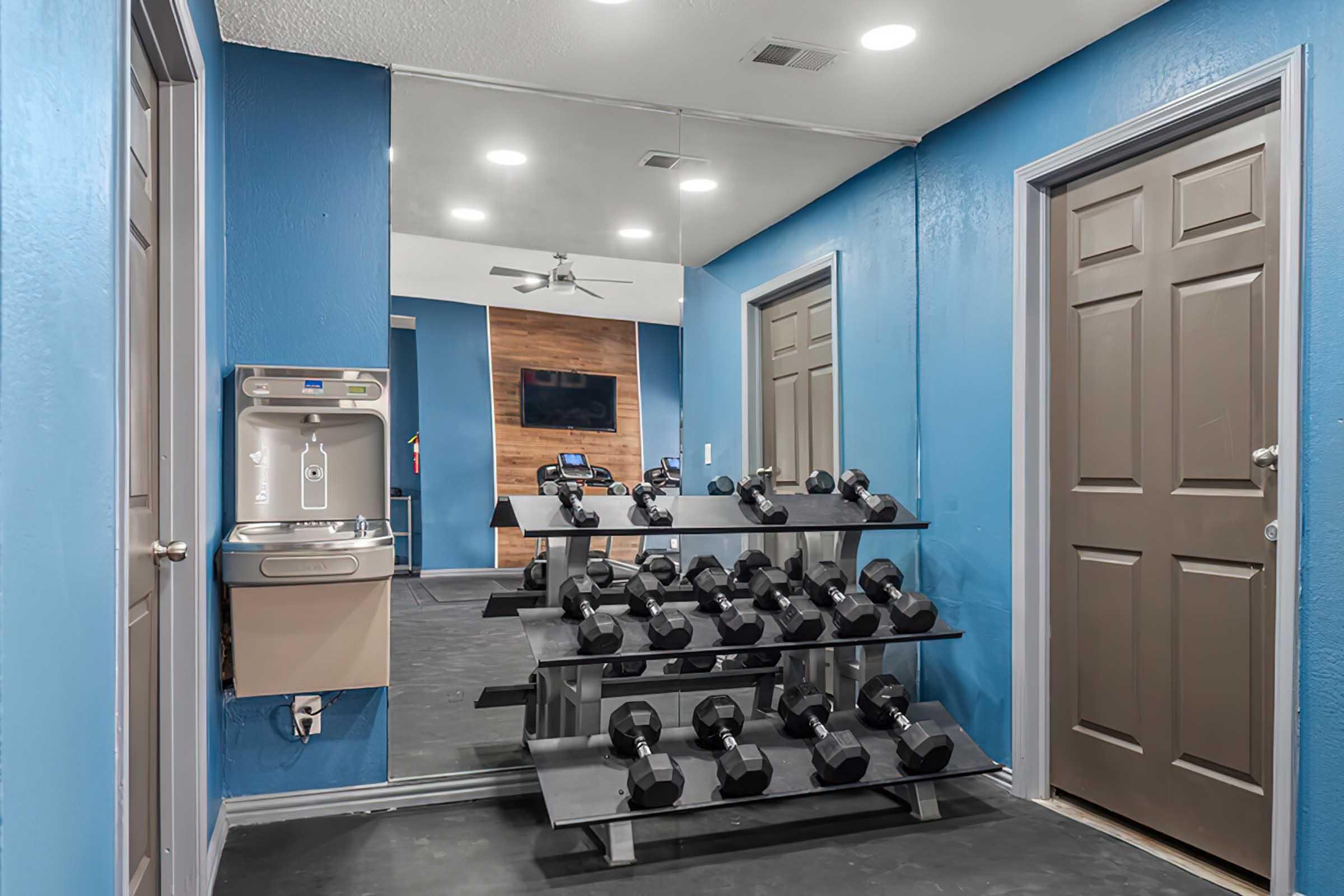 A well-lit fitness room featuring a rack of dumbbells, a water fountain, and mirrored walls. The room has blue walls and multiple doorways, creating a spacious workout environment.