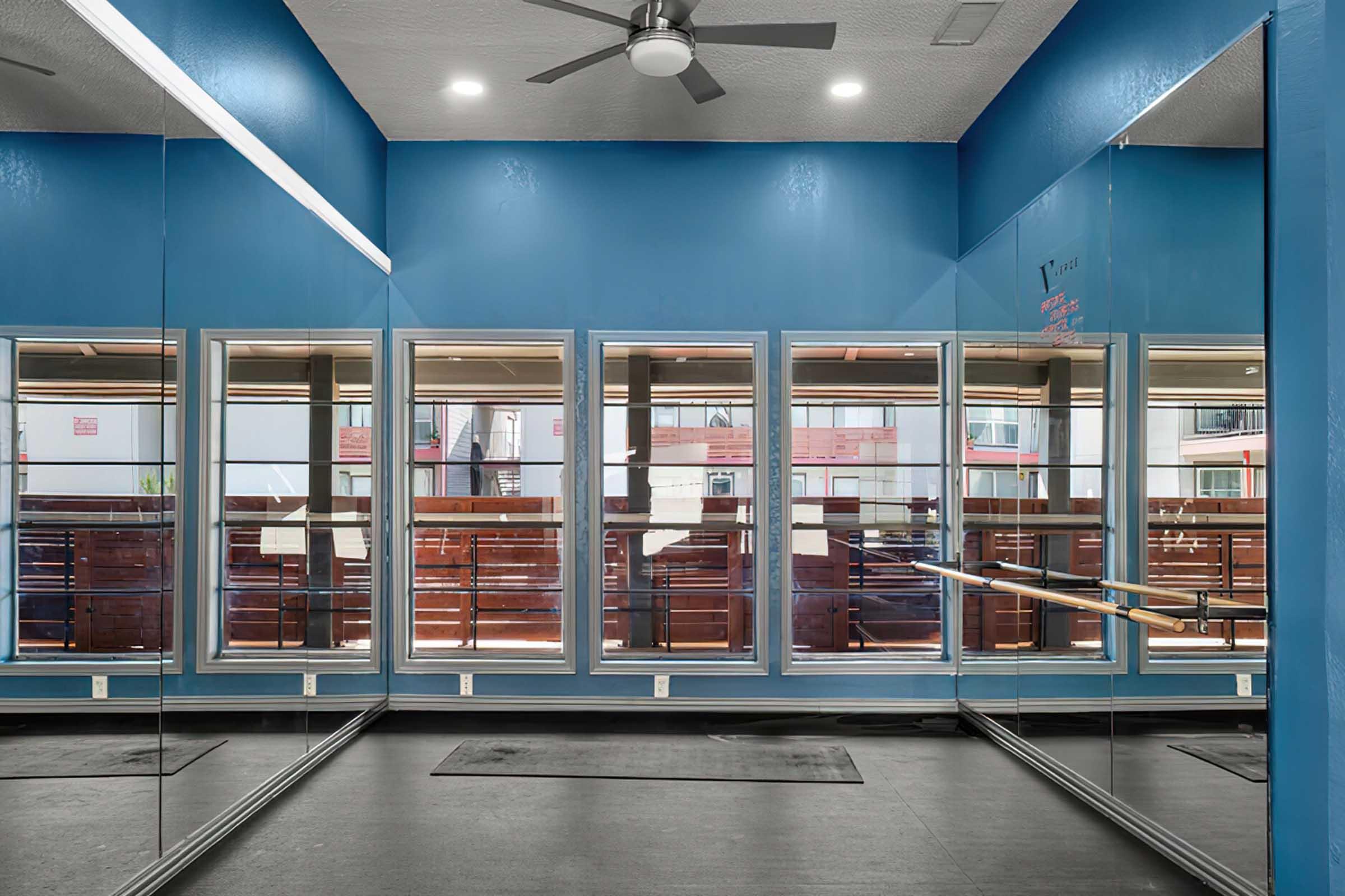 A bright, airy studio featuring blue walls and large windows. Mirrors line the walls, reflecting the space. The floor is dark, with a few mats visible. Natural light streams in through the windows, creating a welcoming environment, likely intended for dance or fitness activities.