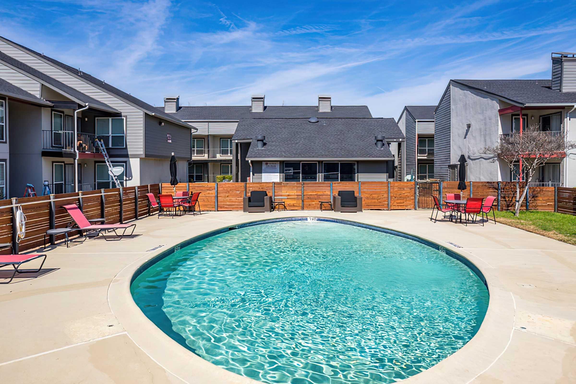A sunny outdoor swimming pool surrounded by lounge chairs and a wooden fence, with two buildings in the background featuring balconies. The sky is blue with a few clouds, creating a bright and inviting atmosphere.