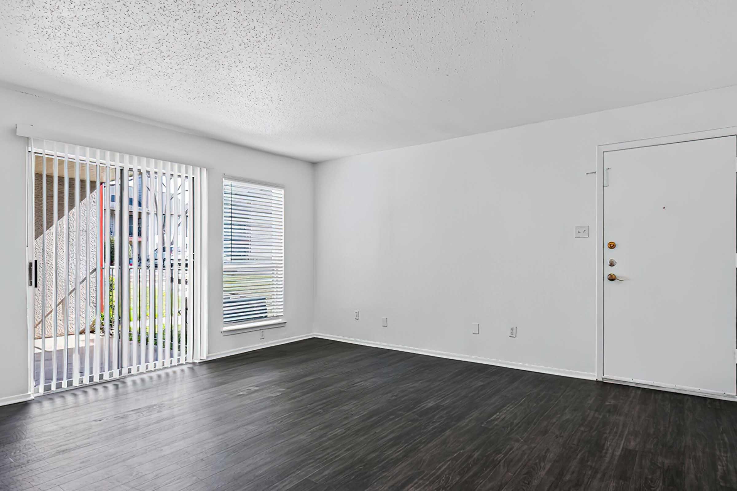A spacious, well-lit room with white walls and a dark hardwood floor. Large windows with blinds allow natural light, and a sliding door opens to the outside. A simple entry door is visible, and the overall ambiance is clean and modern, suitable for living or office space.