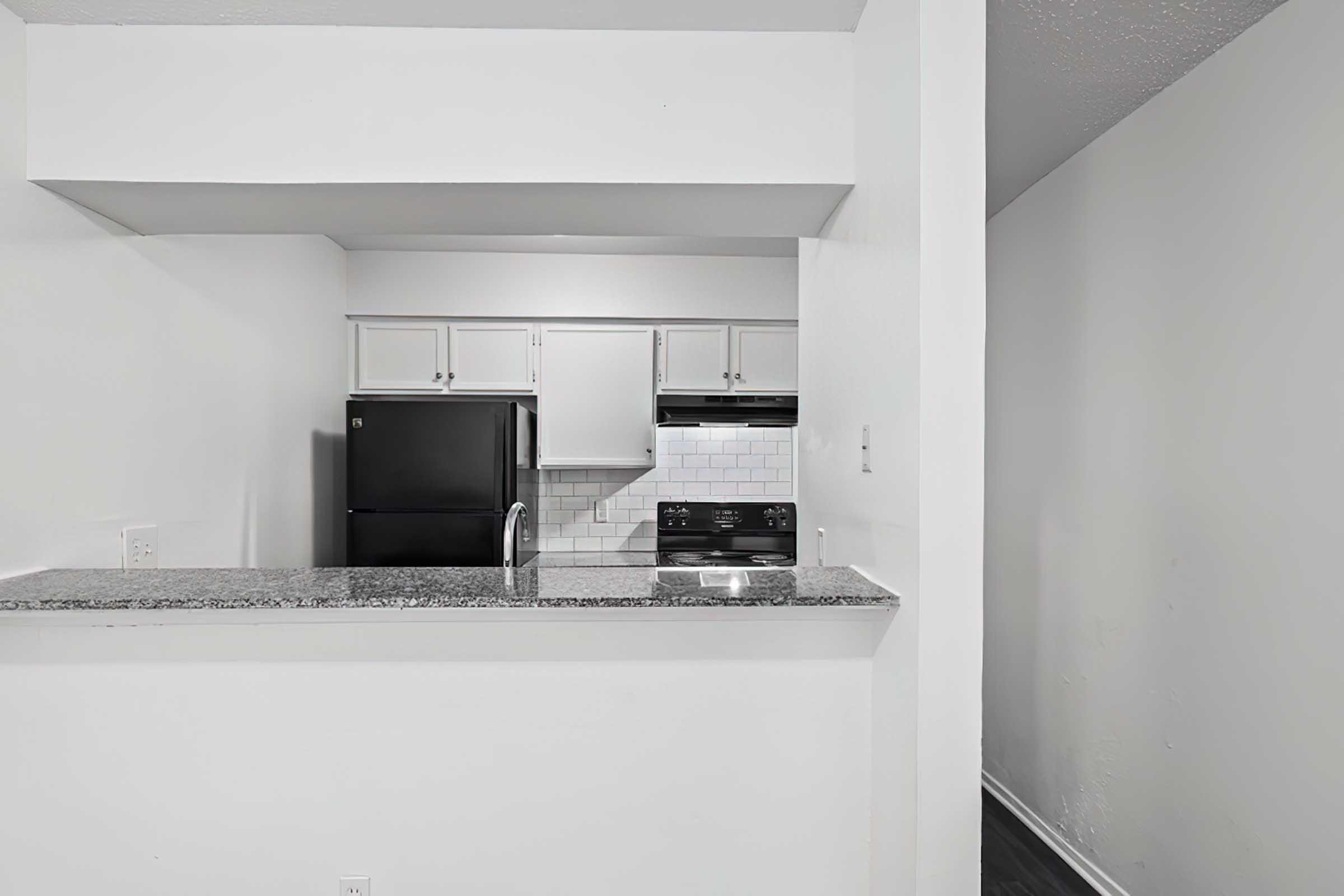 A modern kitchen area with a granite countertop, featuring a black refrigerator, a stove, and white cabinets. The kitchen has a clean, minimalist design with a light-colored wall and an open layout.