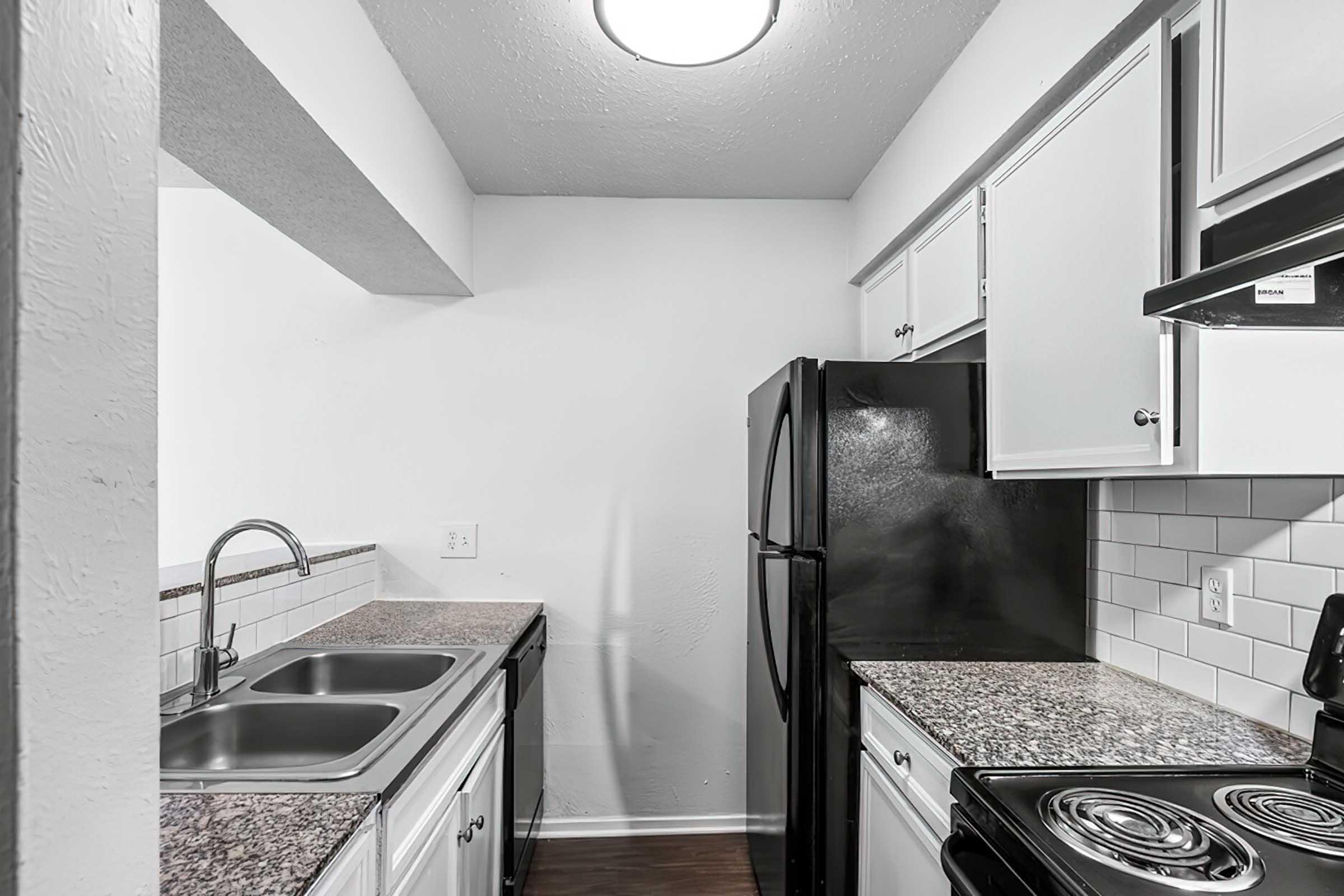 A modern kitchen featuring black appliances, including a refrigerator and stove. The countertops are a speckled granite while the cabinetry is white, providing a bright contrast. There is a double sink with a polished chrome faucet, and the walls are painted a soft light color, creating a clean and contemporary atmosphere.