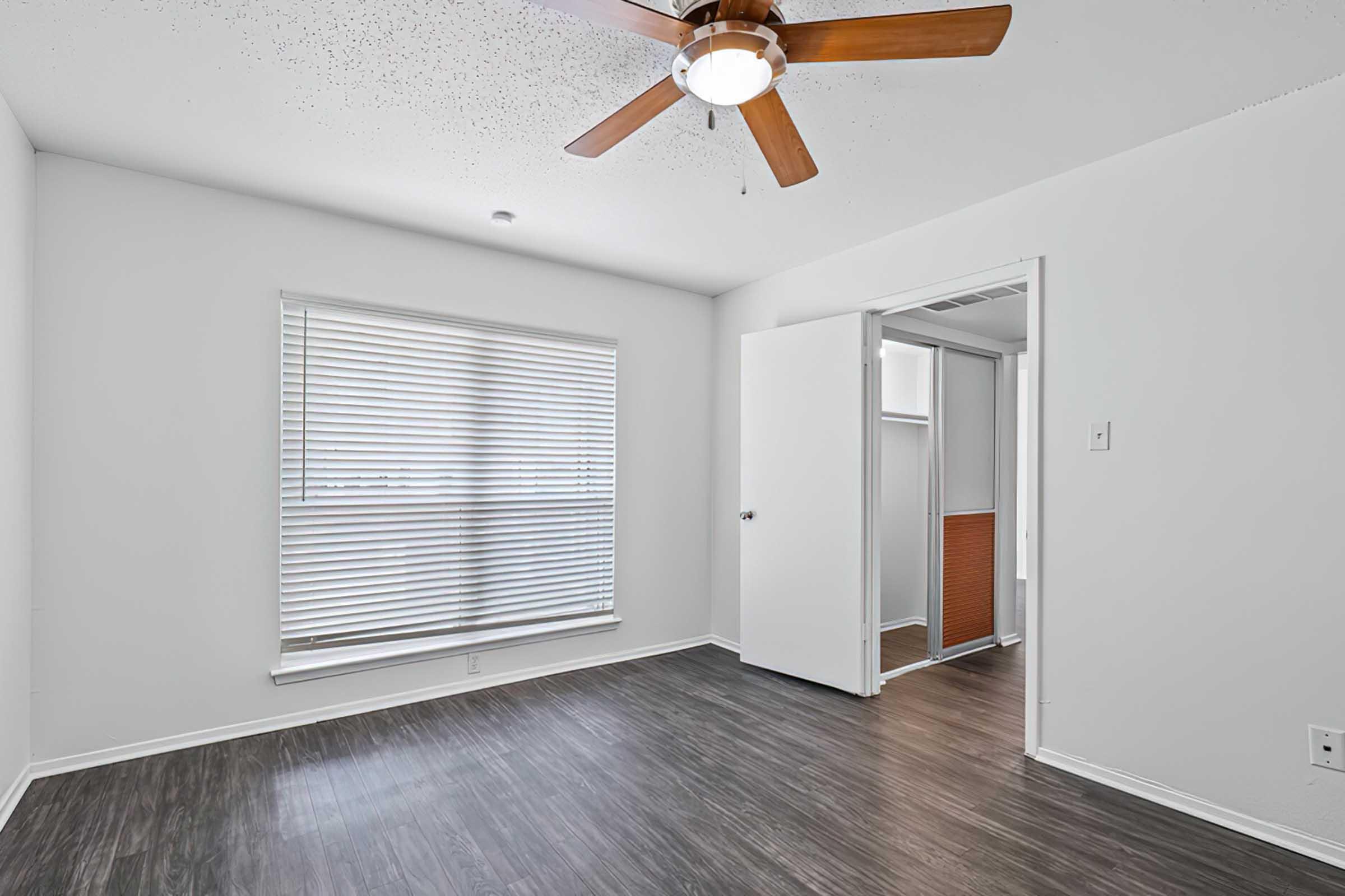 A bright, empty room featuring a ceiling fan and a window with blinds. The walls are painted white, and the floor has a dark, wood-like finish. A partially open closet is visible on one side, and the overall atmosphere is clean and spacious.