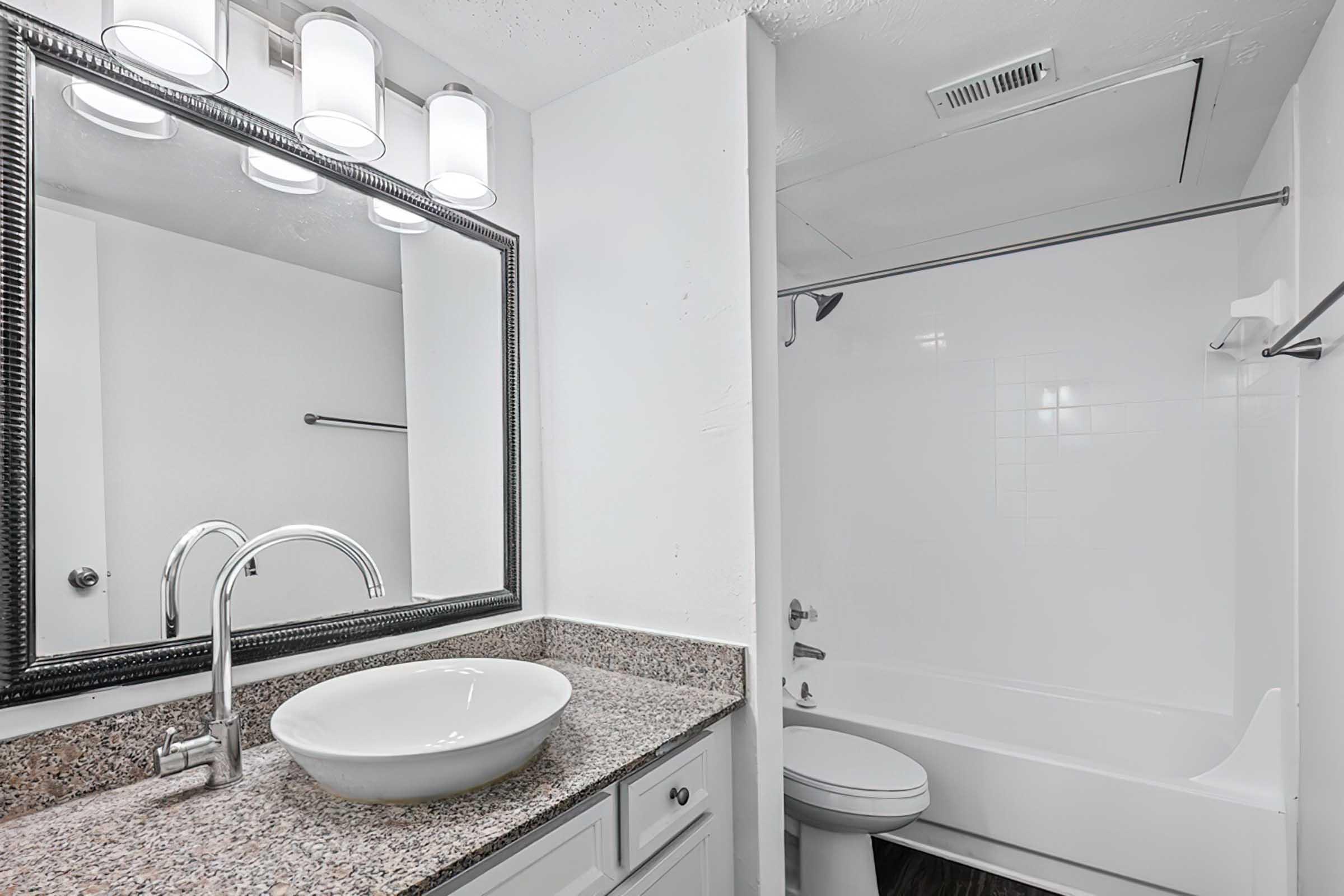 Modern bathroom featuring a sleek vanity with a white vessel sink, granite countertop, and a large framed mirror. The space includes a bathtub with a shower rod, white tiled walls, and a clean, minimalist design. Bright lighting from above enhances the airy feel.