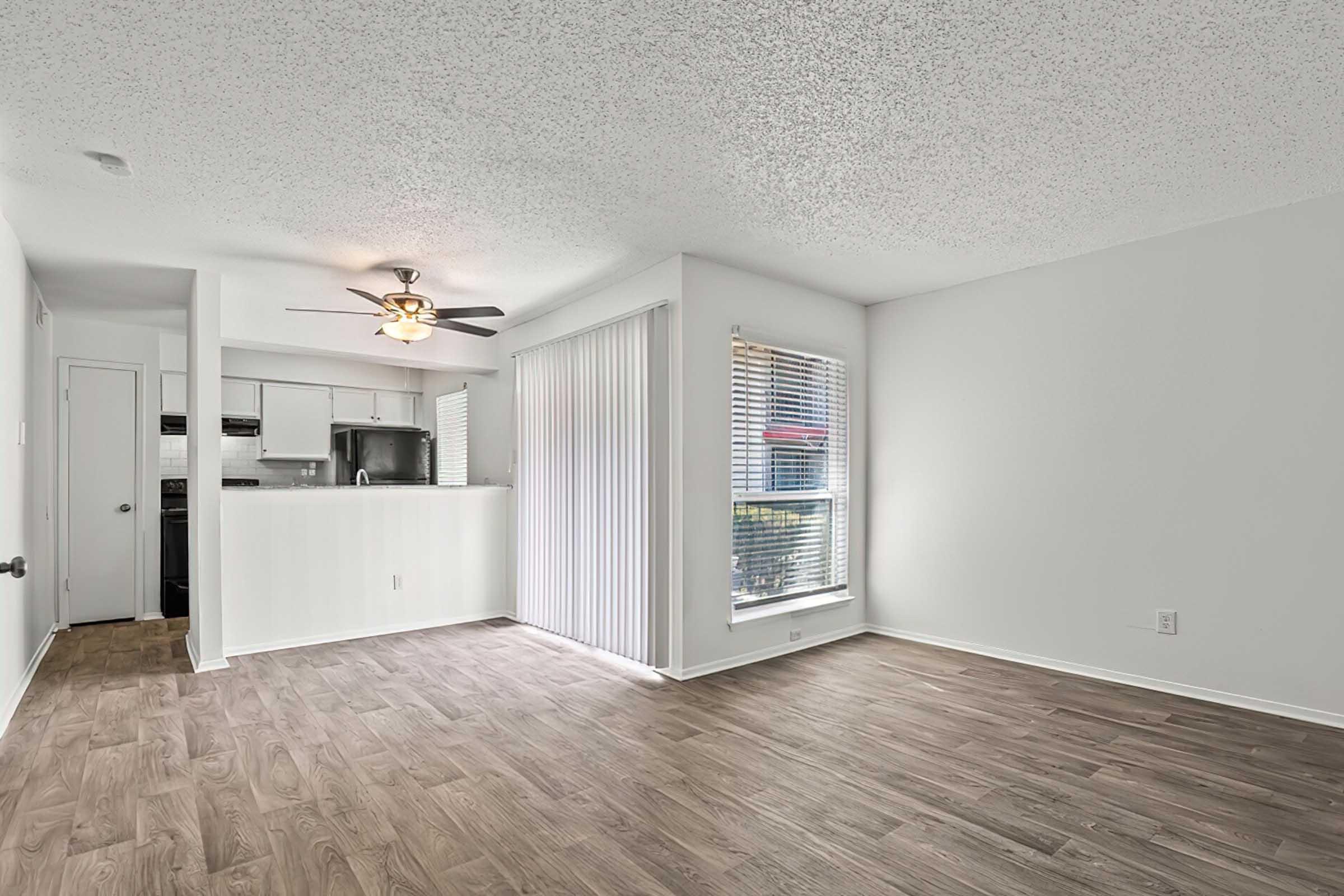 Spacious, bright living area with wood-style flooring, featuring a ceiling fan and large windows. Open layout leads to a modern kitchen with sleek cabinets. Sliding glass door provides access to outdoor space. Light-colored walls create an airy ambiance. Ideal for relaxation and entertaining.
