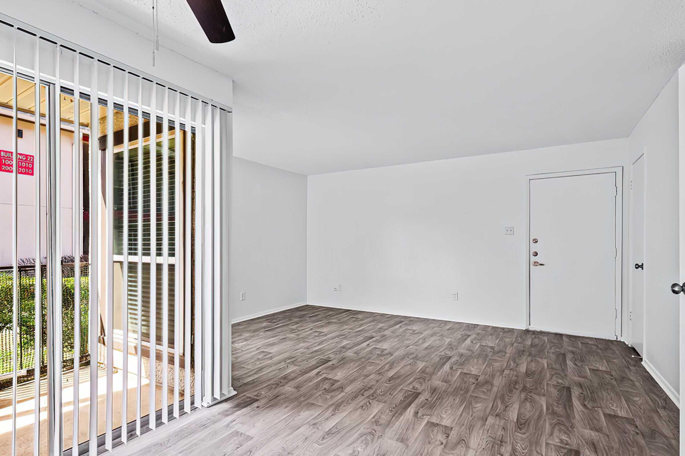 A bright and empty living room with light-colored walls and a smooth wooden floor. Large windows with vertical blinds offer natural light, and a doorway leads to an exterior balcony. A ceiling fan is visible, enhancing the airy feel of the space.