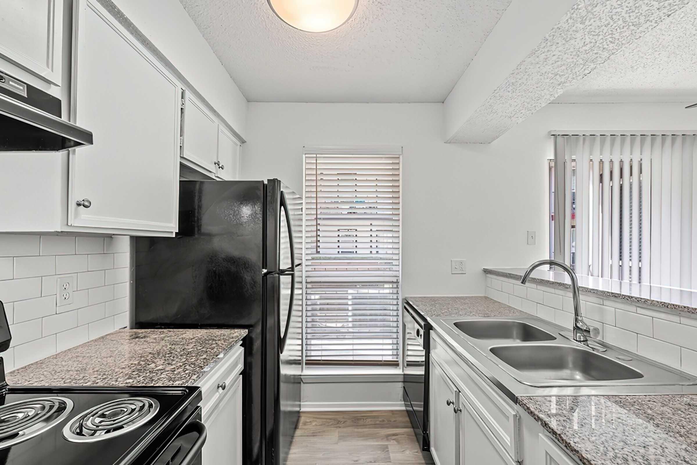 Modern kitchen with white cabinetry, black appliances, and granite countertops. Features a black refrigerator, stove, and a double sink under a window with blinds. Bright atmosphere with light-colored walls and a neutral floor.