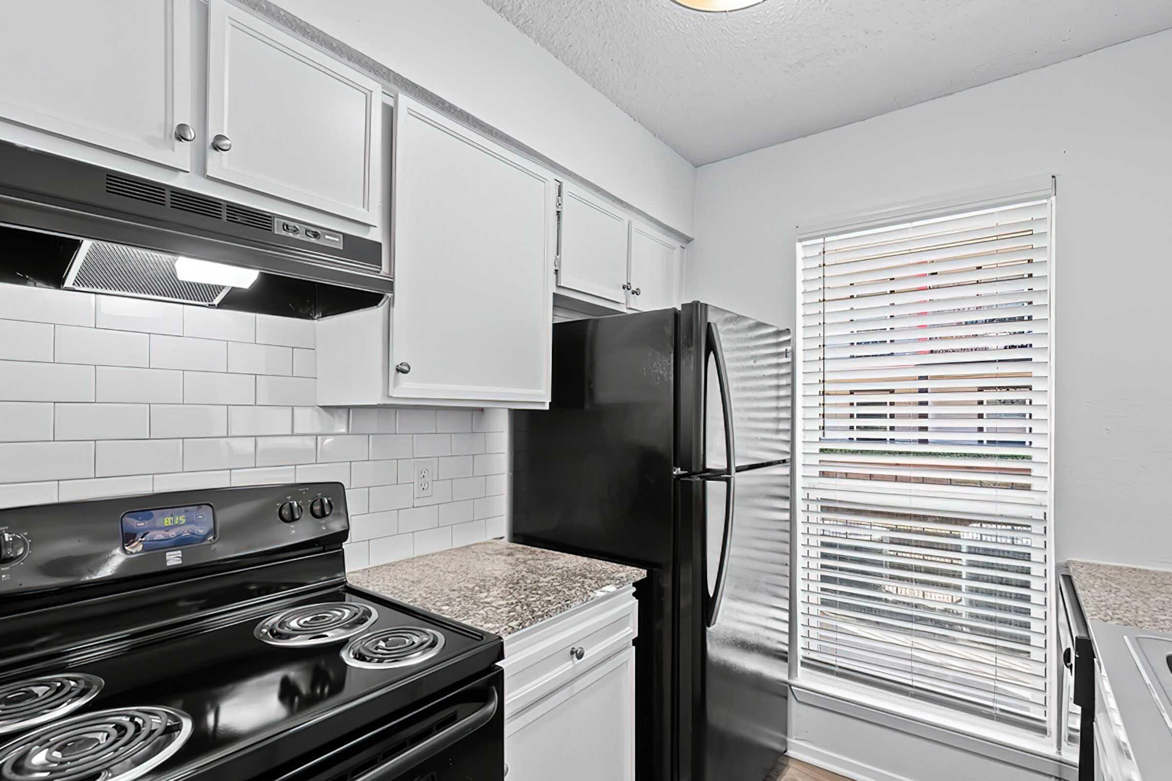 Modern kitchen featuring a black refrigerator and stove, white cabinets, and a granite countertop. There are white tile backsplashes and a window with blinds allowing natural light in. The overall design is clean and contemporary, highlighting efficient use of space.