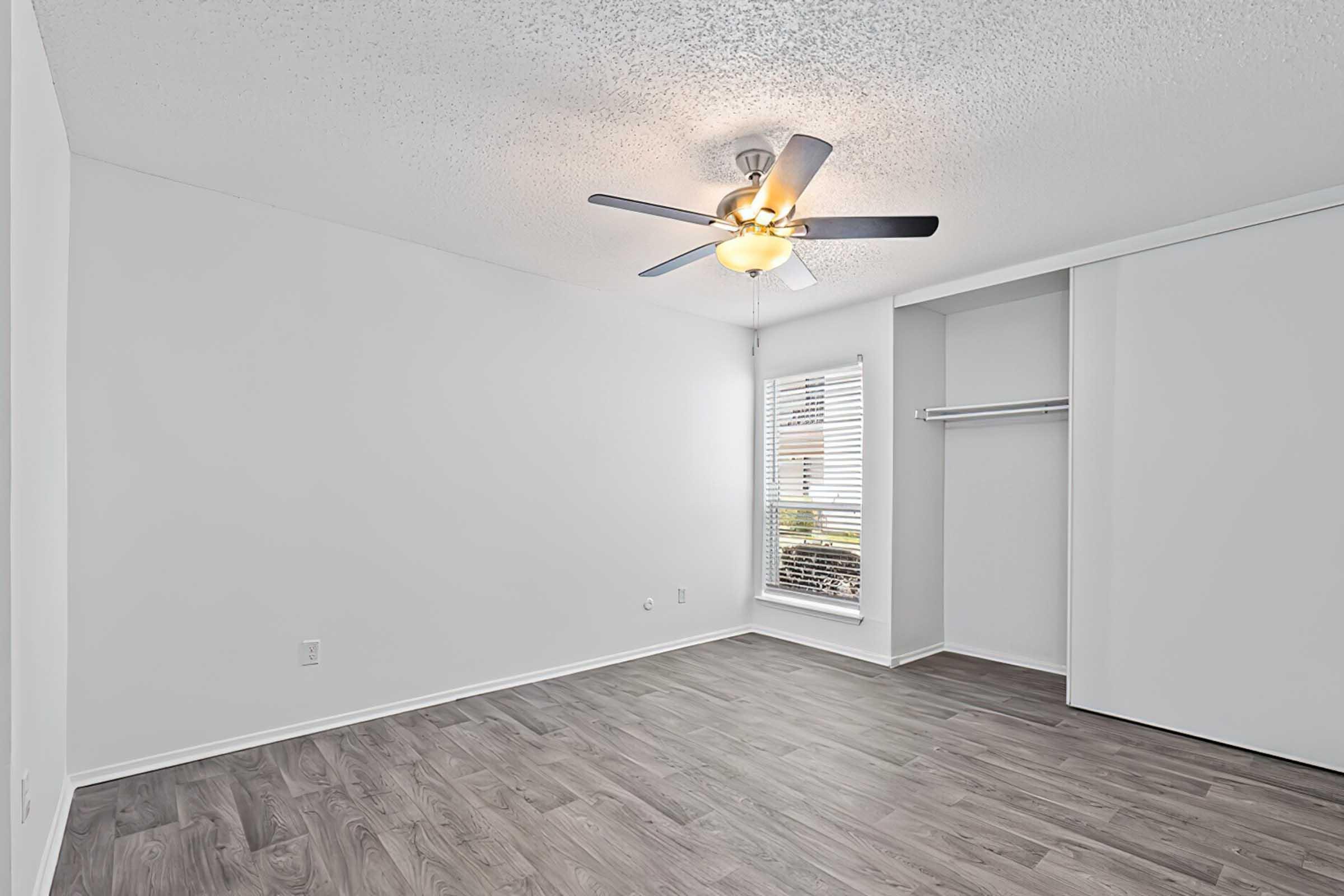 Empty room with light gray walls and a ceiling fan. The floor is covered in laminate wood flooring. A window with blinds allows natural light to enter, and a closet is visible in the corner. The space appears modern and inviting, suitable for a variety of furnishings.