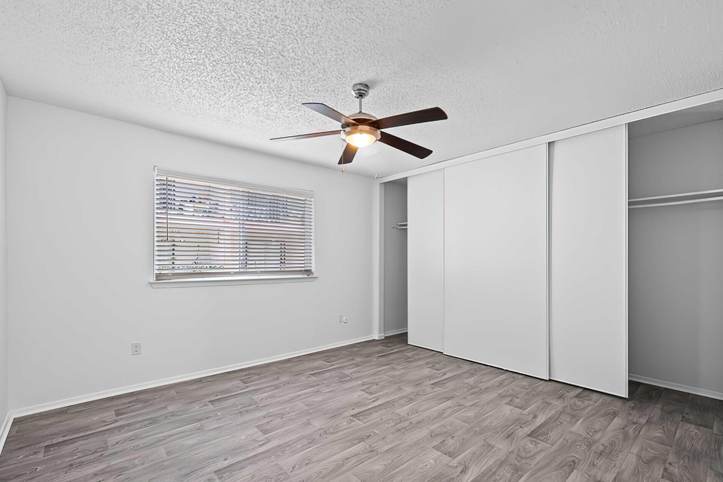 A well-lit, empty bedroom featuring light gray walls, a ceiling fan with wooden blades, a window with blinds, and light wooden flooring. There is a closet with sliding white doors to the right, creating a clean and modern aesthetic.