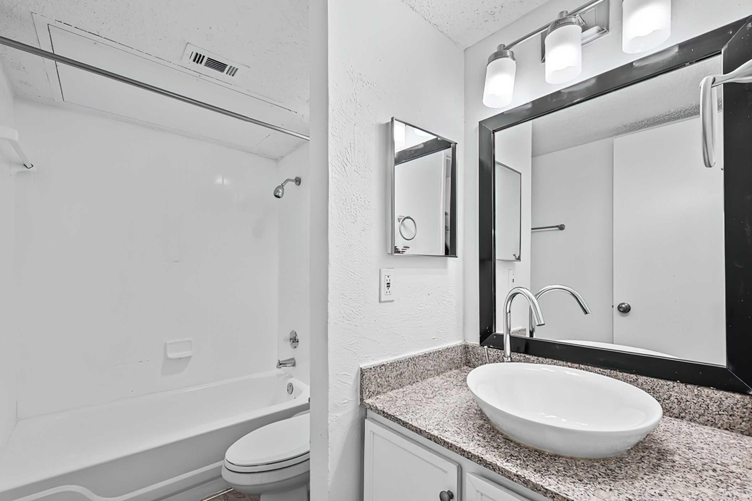 A modern bathroom featuring a white bathtub with a shower, a sleek, round sink atop a granite countertop, a rectangular mirror, and a light fixture with three bulbs. The walls are light-colored, and the overall design is clean and contemporary.