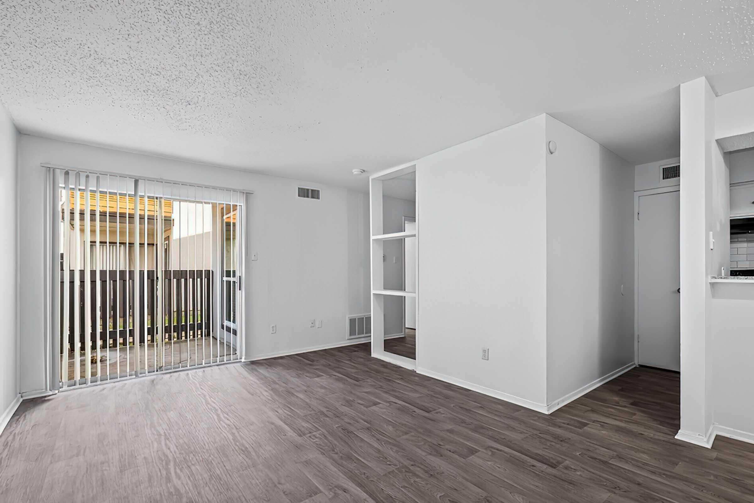 A spacious living room featuring light-colored walls and hardwood-style flooring. Large sliding glass doors lead to a small outdoor area. A mirror-fronted closet is visible in the corner, and there's an open layout connecting to a kitchen area. The overall atmosphere is bright and airy.
