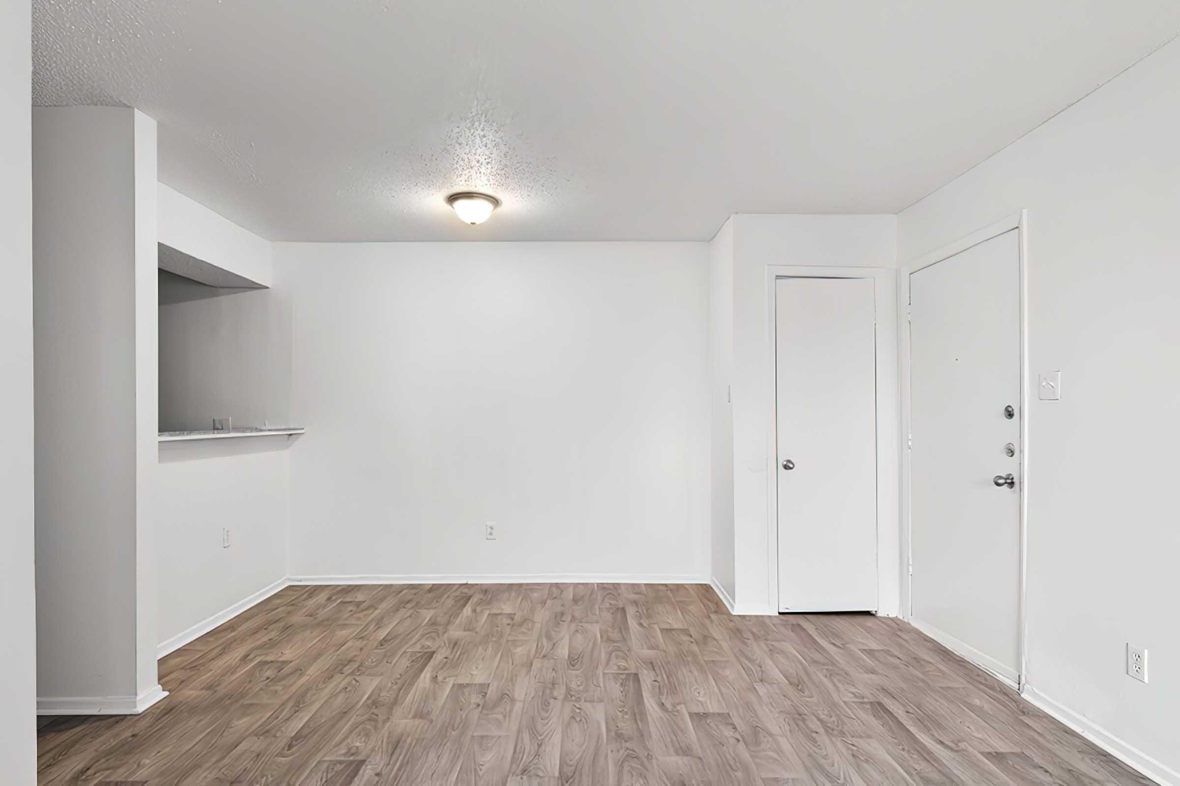 A spacious, unfurnished room with light-colored walls and a wood-style floor. There is a ceiling light fixture, a small opening leading to another room, and a door on the right side. The overall ambiance is bright and minimalistic, suitable for various interior designs.