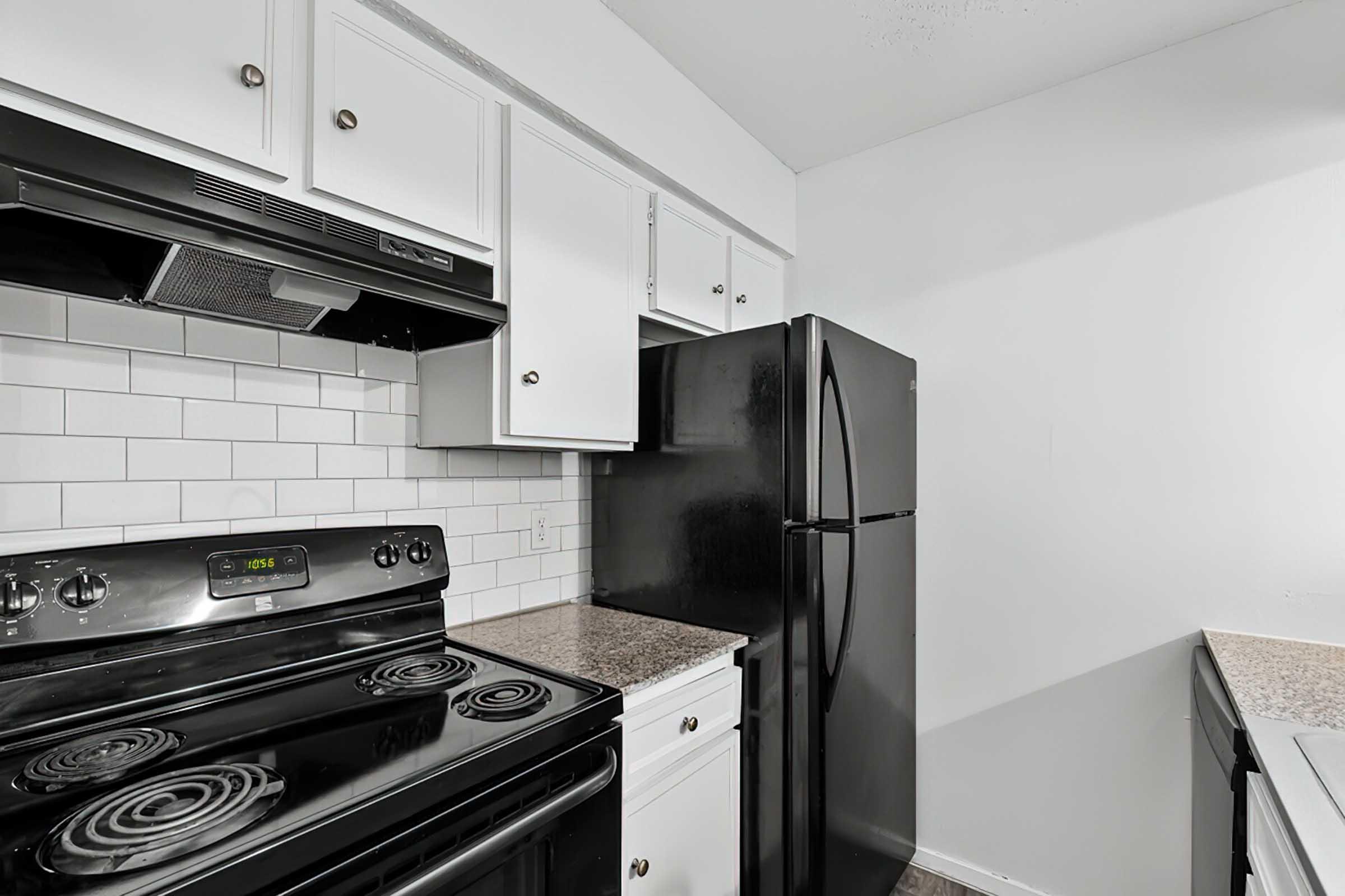 A modern kitchen featuring black appliances, including a stove, refrigerator, and dishwasher. The cabinets are white, and the backsplash consists of white subway tiles. The countertop is a light speckled pattern, providing a clean and functional cooking space. The overall color scheme is black and white.
