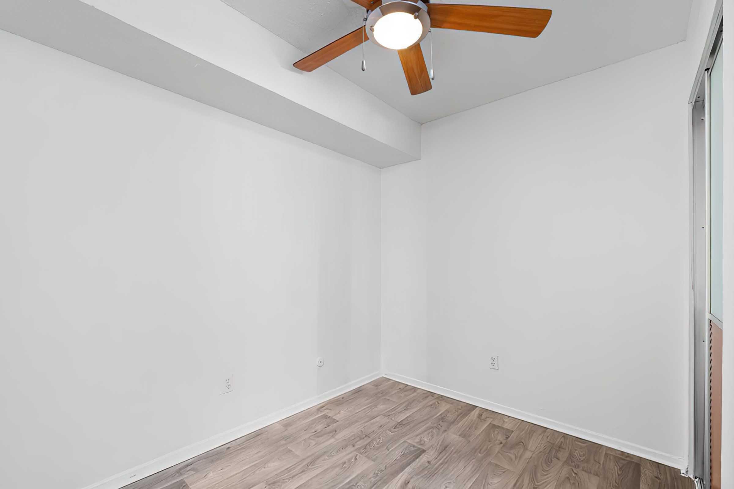 Empty room with white walls and a wood-paneled floor. A ceiling fan with wooden blades is mounted on the ceiling, and there are no furnishings or decorations in the space. Natural light enters from a window, contributing to a bright and airy ambiance.