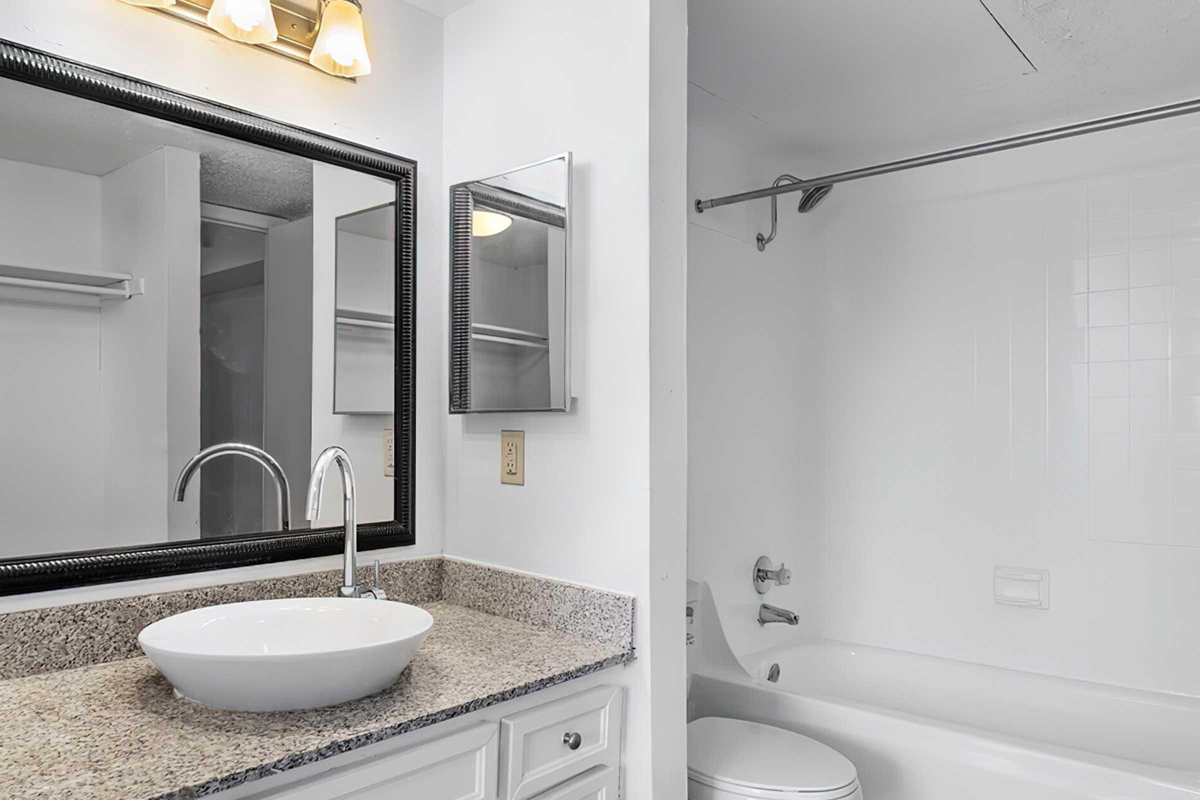 A clean, modern bathroom featuring a white sink with a circular bowl atop a granite countertop, a large mirror above the sink, and a bathtub with a shower curtain. The walls are painted in a light color, and there are minimal fixtures to maintain a spacious feel.