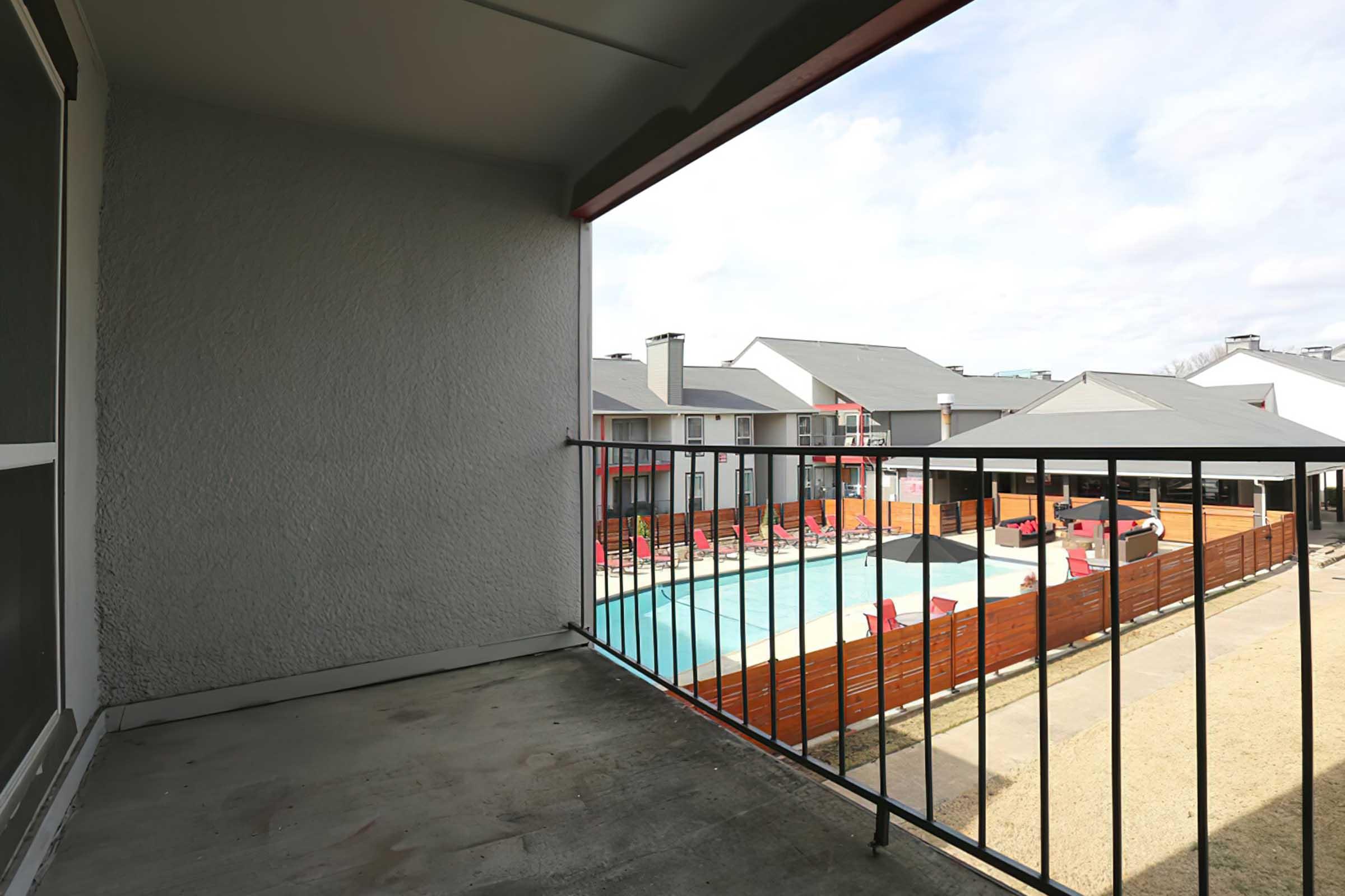 A view from a balcony showing a fenced pool area surrounded by lounge chairs and a grassy lawn. In the background, there are several apartment buildings with gray roofs and a partly cloudy sky.