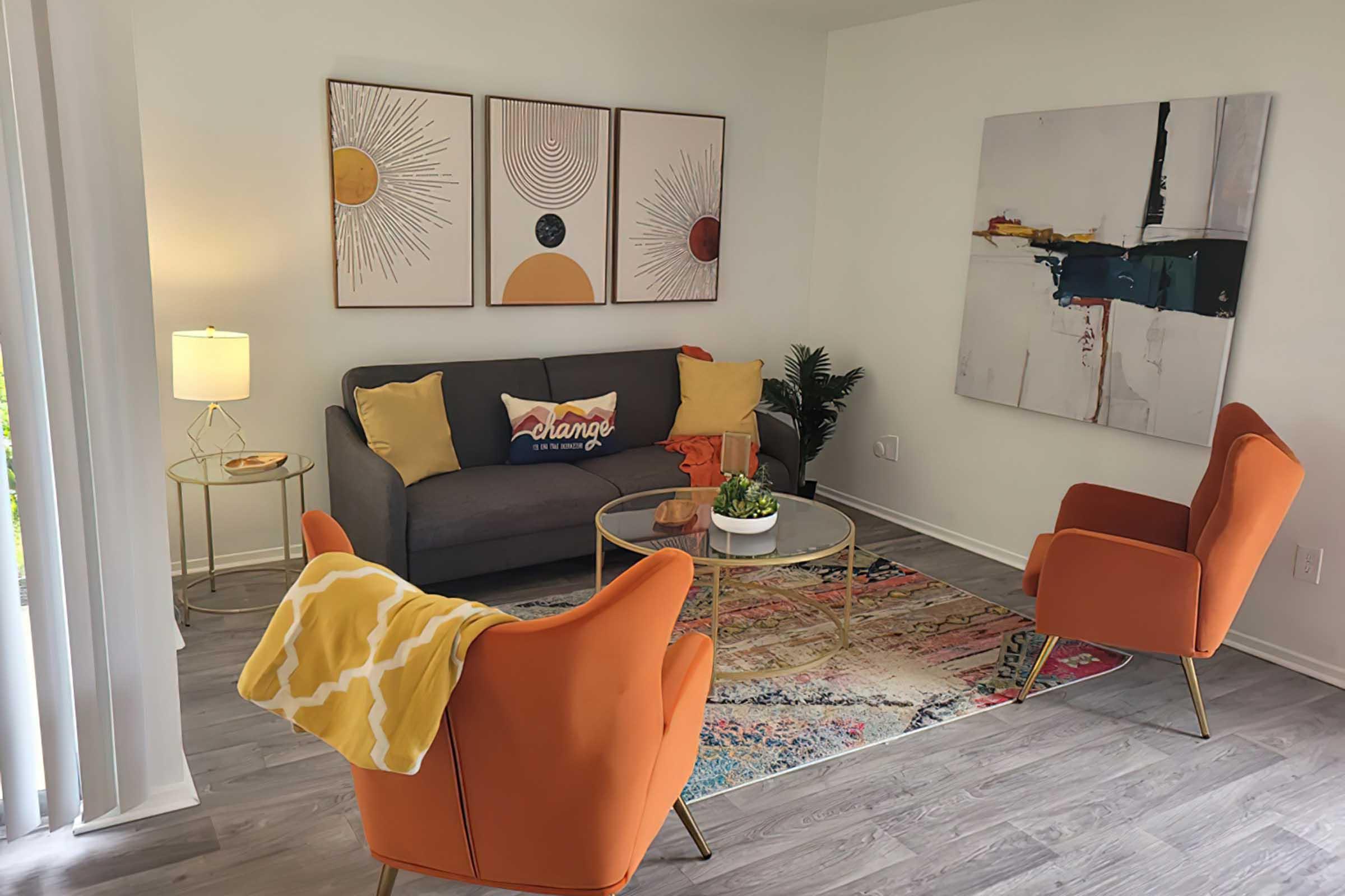 A modern living room featuring a gray sofa with colorful cushions, two orange accent chairs, and a round coffee table. The walls are adorned with abstract art and three framed prints. A lamp and a small plant add to the decor, while a vibrant area rug ties the space together. Natural light fills the room.
