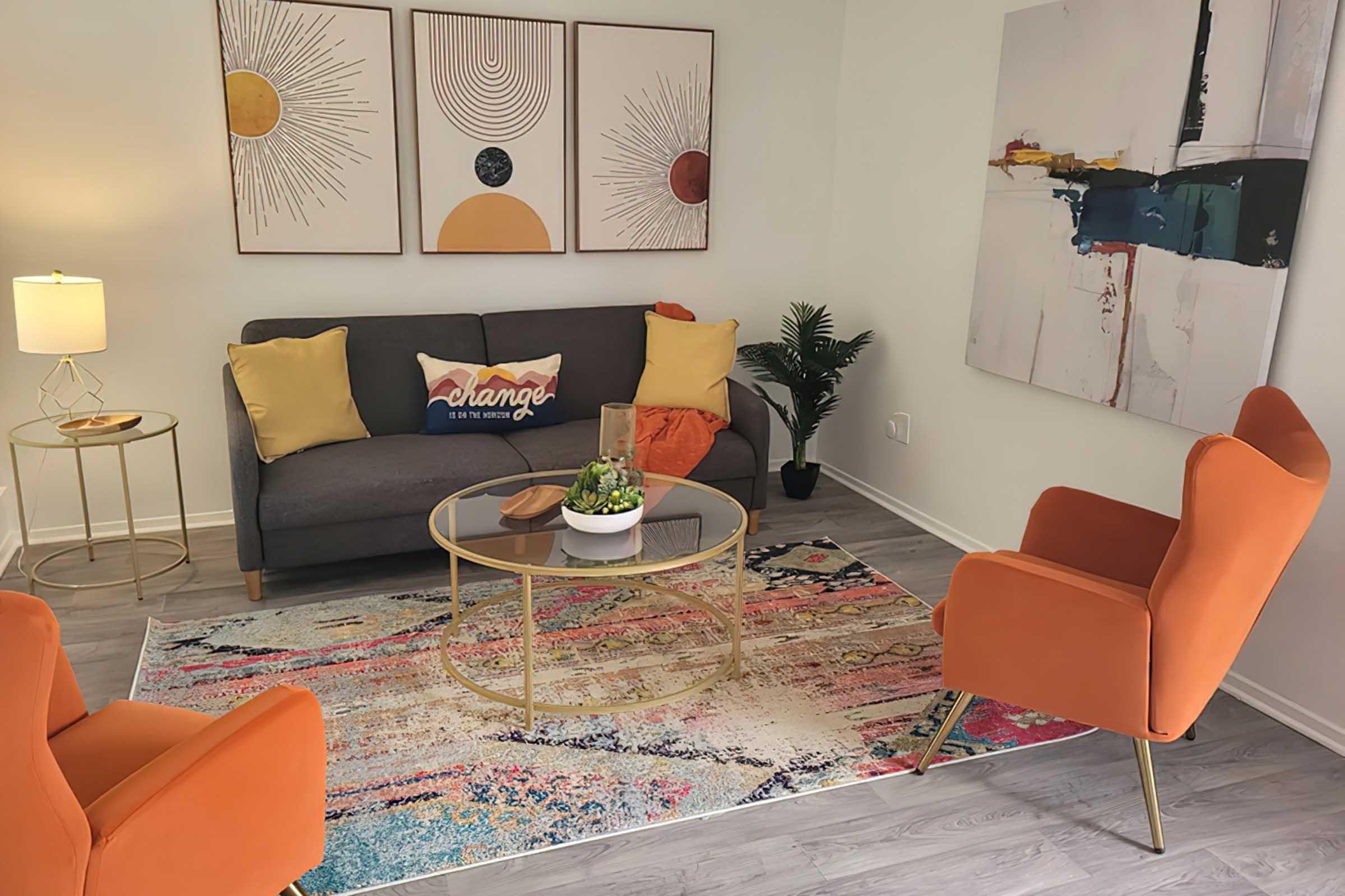A modern living room featuring a gray sofa with colorful pillows, two orange armchairs, a round glass coffee table, and a decorative plant. The floor is covered with a vibrant area rug, and three framed wall art pieces are hung above the sofa. A lamp adds warm lighting to the space.